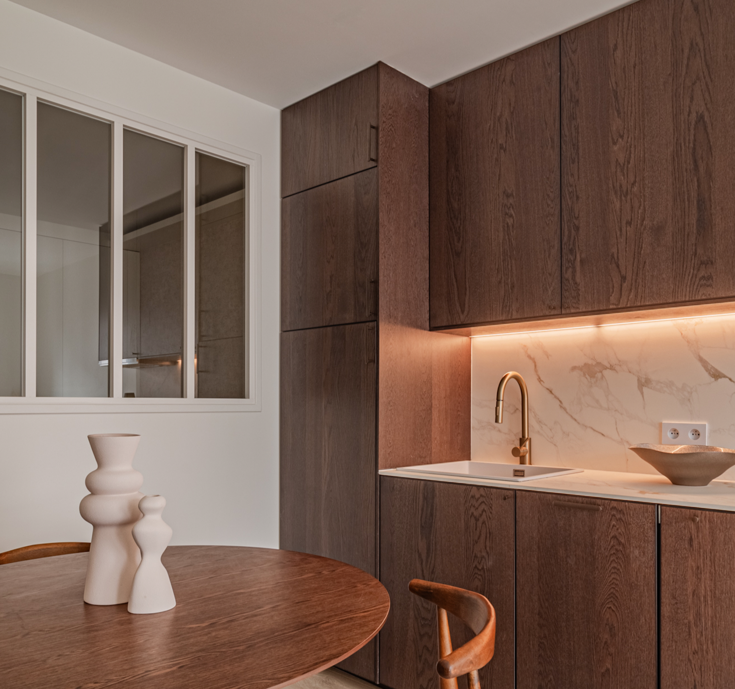 A modern kitchen corner featuring wooden cabinetry, a marble backsplash, a gold faucet, a wooden dining table with decorative vases, and a window with white framing.