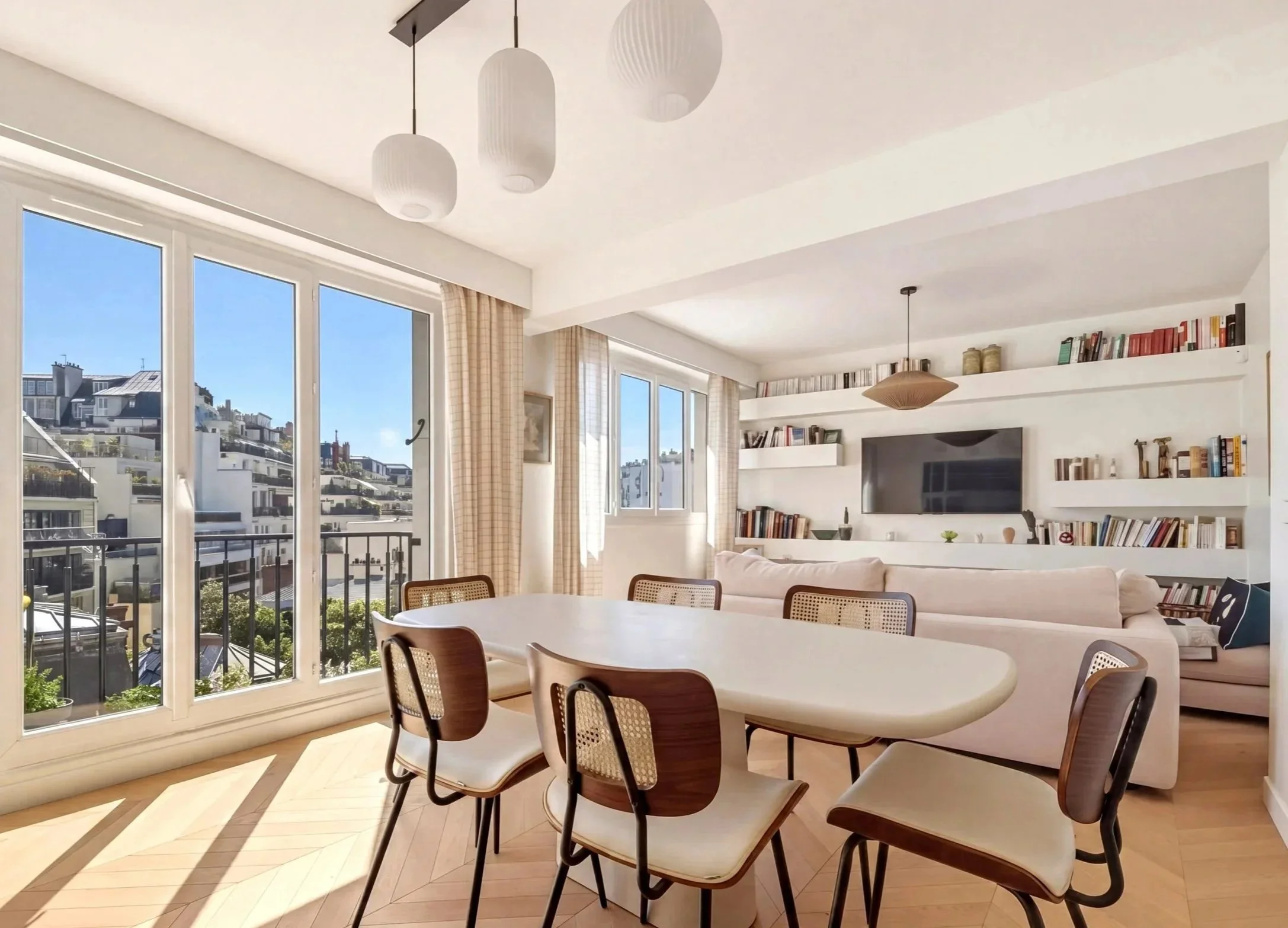 Bright living room with large windows, white walls, wooden flooring, a dining table with chairs, a beige sofa, white shelves with books, and ceiling lamps.