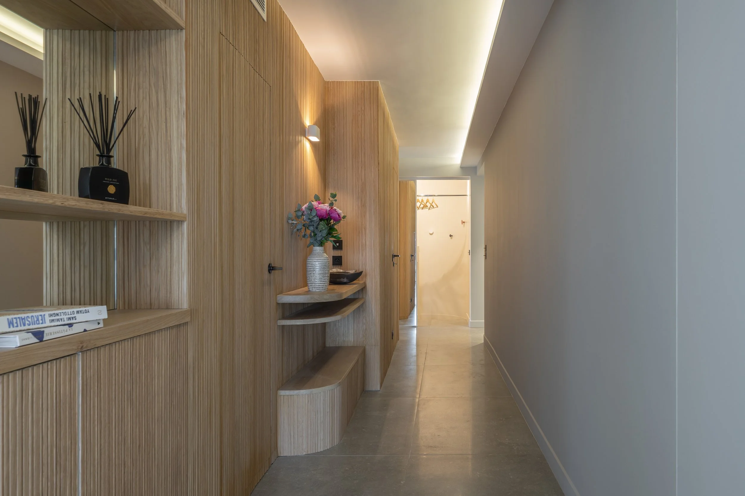 A modern hallway with wooden paneling on one side, a white wall on the other, and a ceiling with built-in lighting. There is a vase with pink flowers on a wooden shelf and two black decorative bottles with reed diffusers. In the background, there is a door and a glimpse of an open closet with hangers.