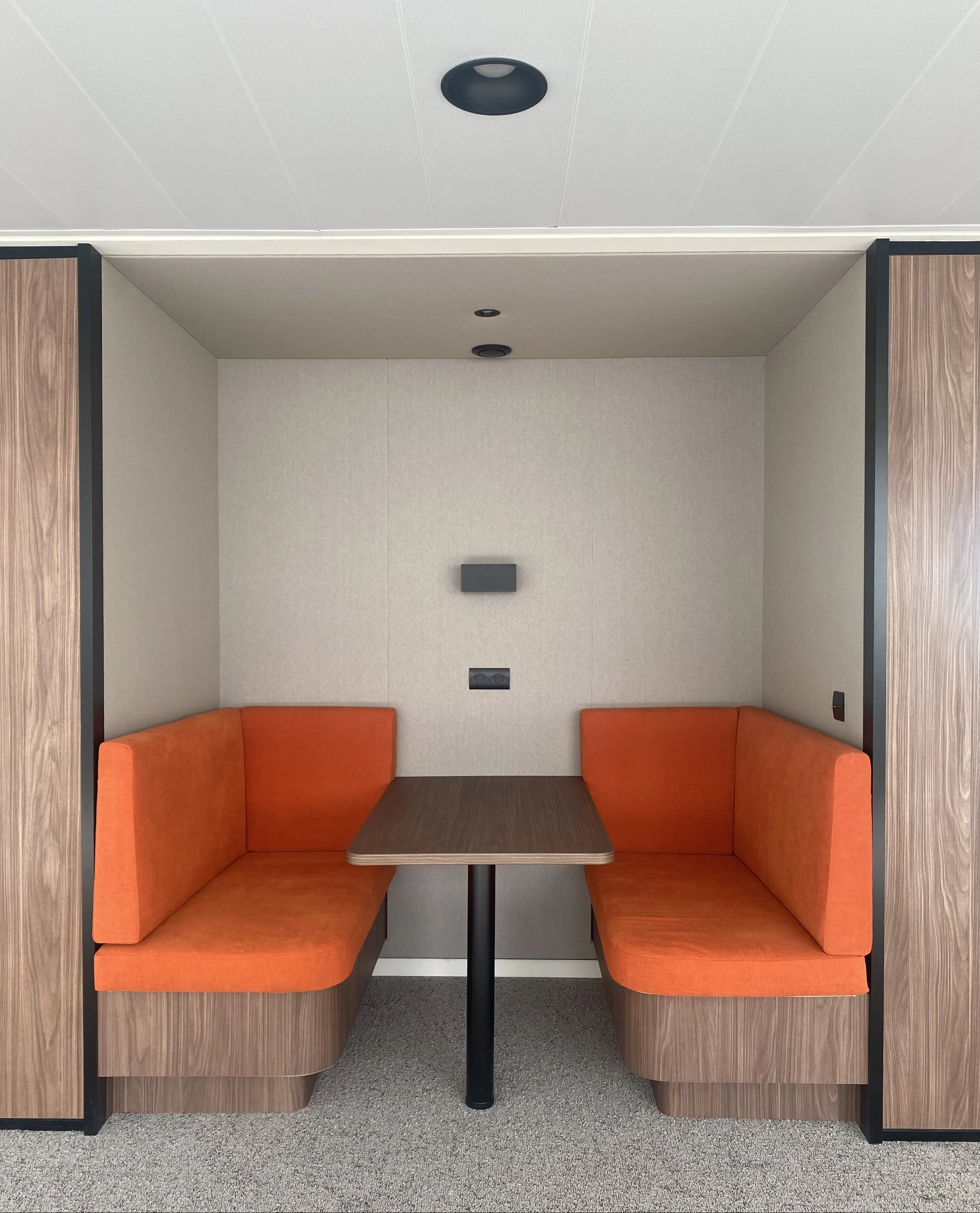 A booth seating area with orange upholstered cushions in a modern office or waiting room, with a small rectangular wooden table and textured beige walls.