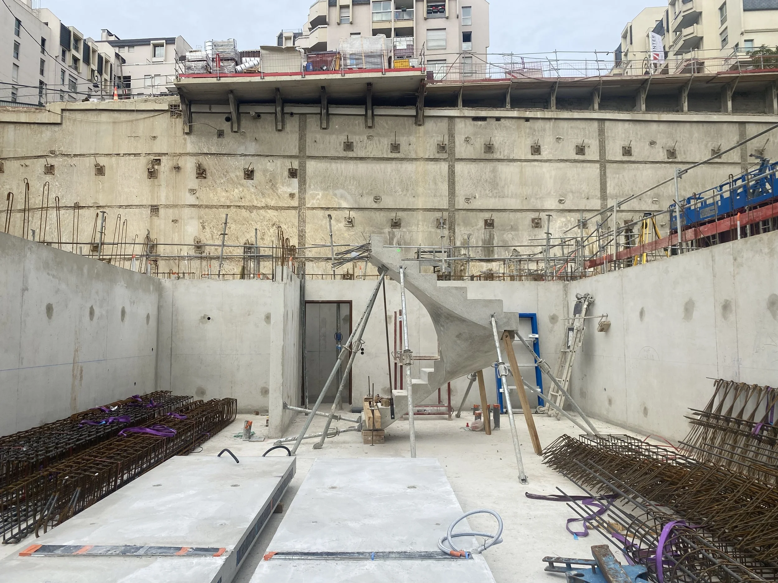 Construction site with unfinished concrete structure, rebar, scaffolding, and building materials. Several buildings are visible in the background.