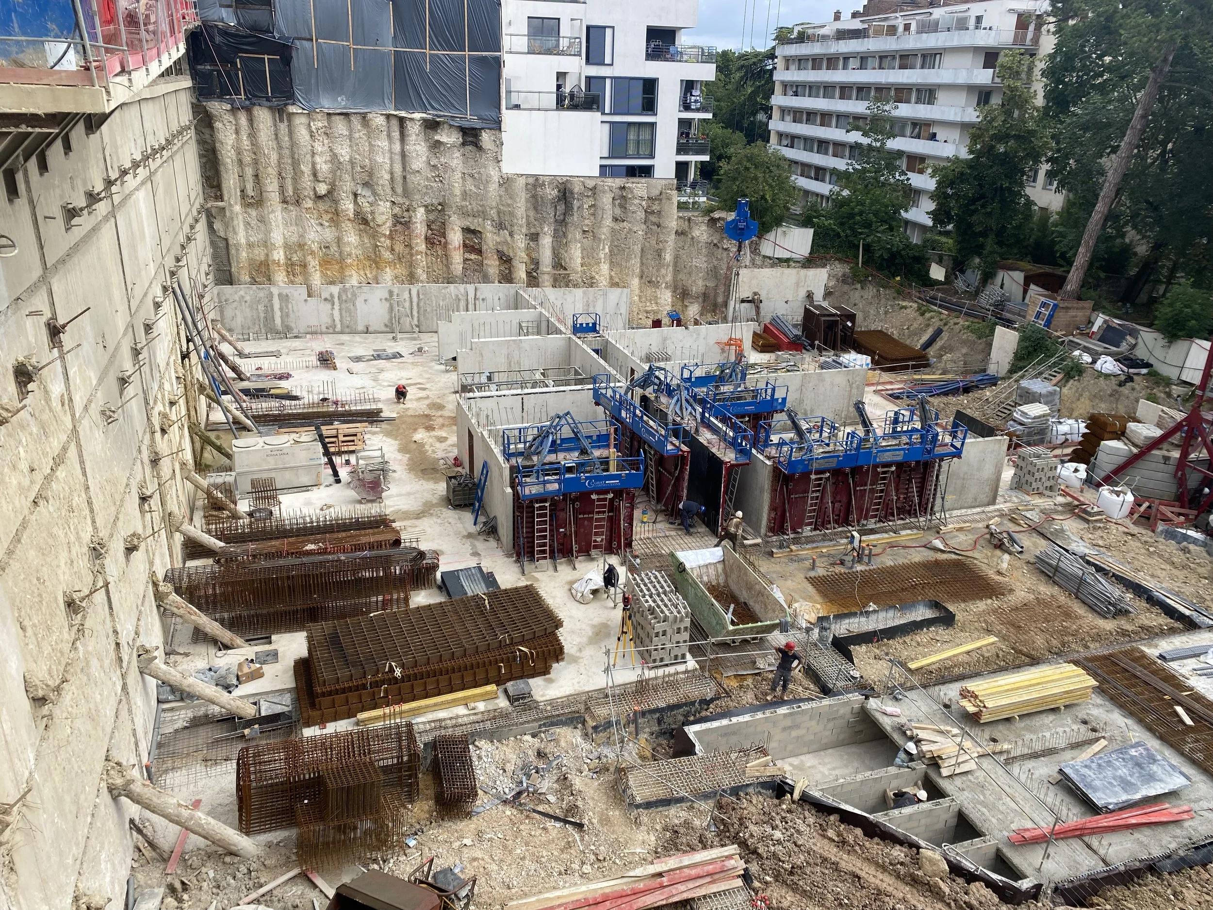 Construction site with workers and equipment at building foundation stage surrounded by residential buildings.