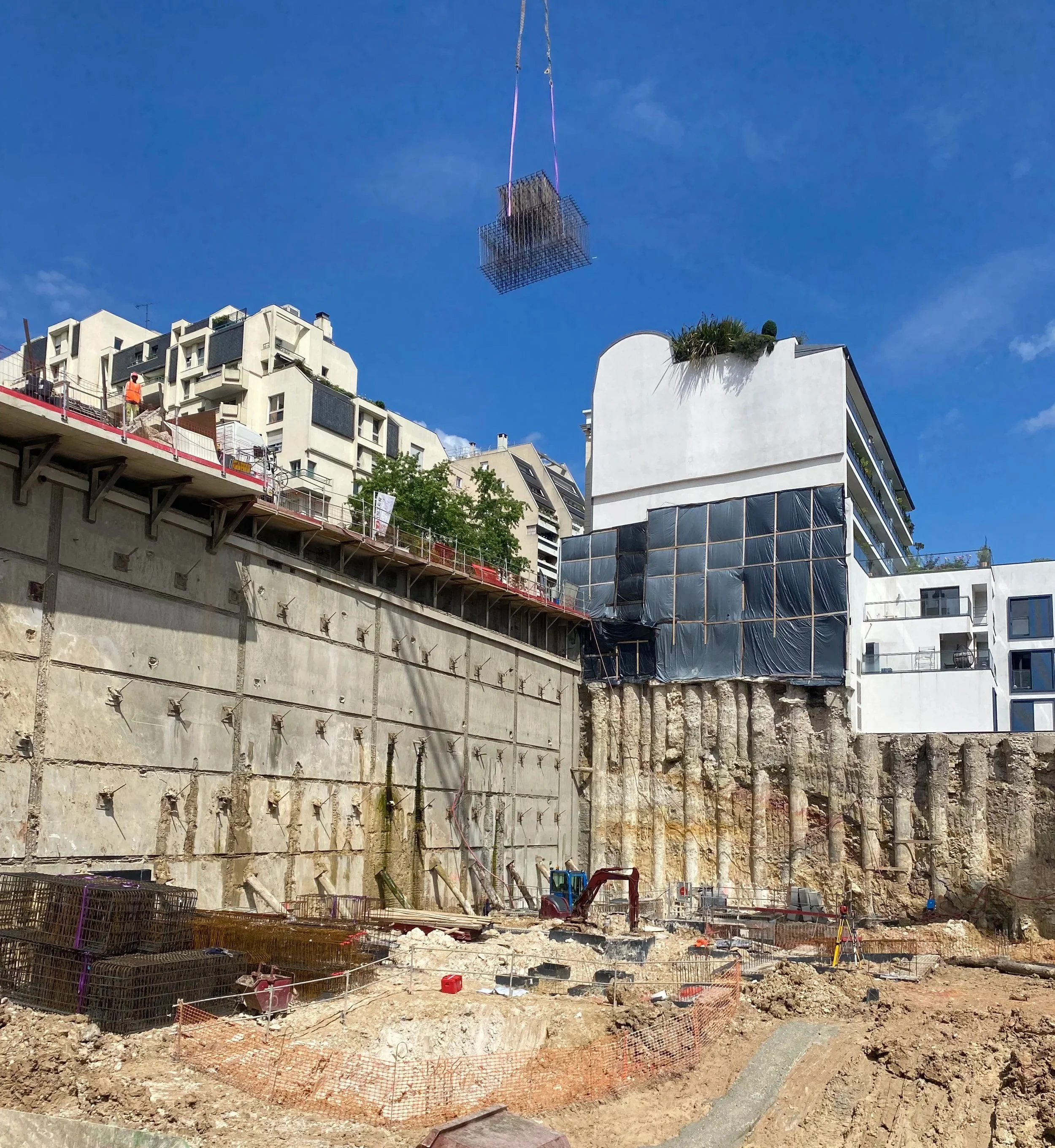 Construction site with building foundation, rebar, safety fencing, and construction workers amid urban buildings and a crane lifting materials into the air.