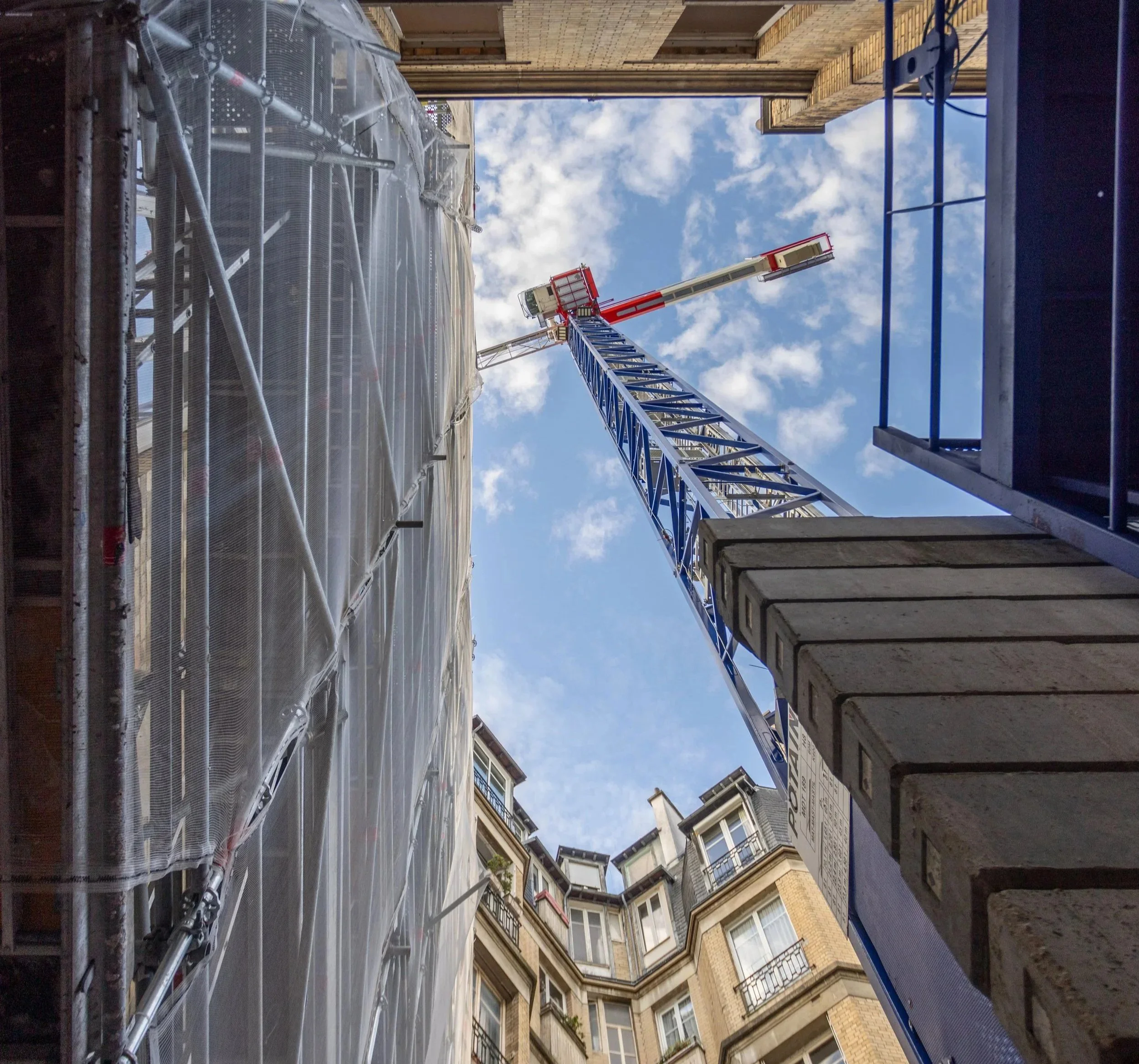 View from the ground looking up at a blue and white construction crane between tall buildings made of brick and concrete under a blue sky with scattered clouds