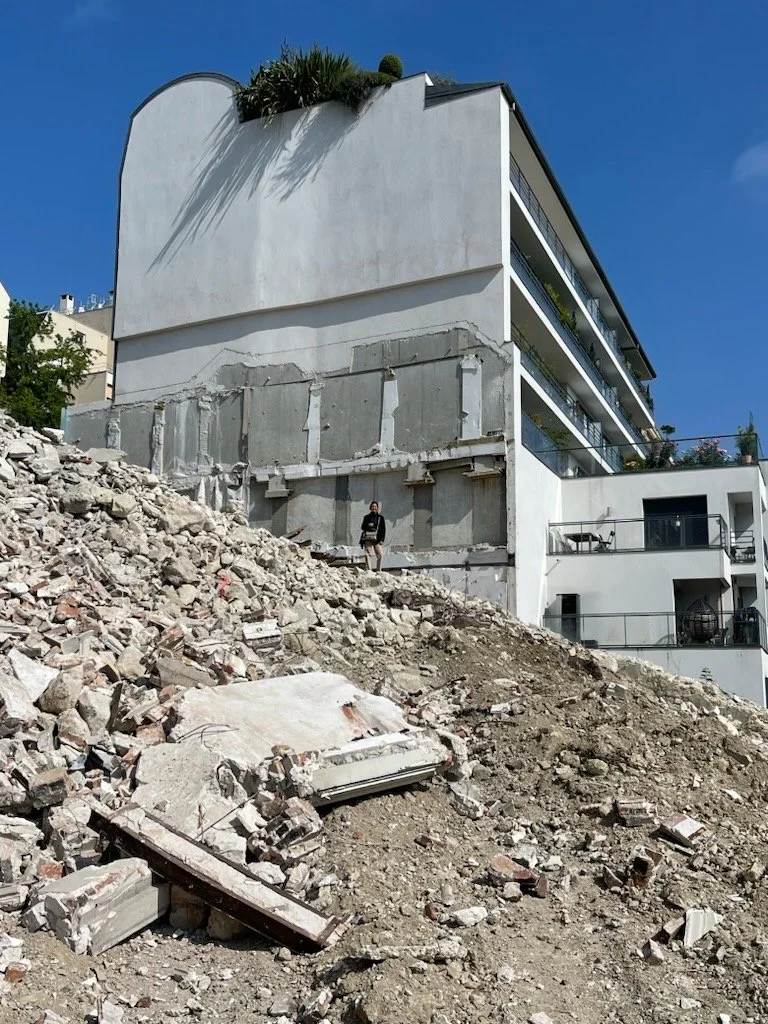 Construction debris and rubble in front of a modern residential building on a hillside, with a woman standing among the rubble.