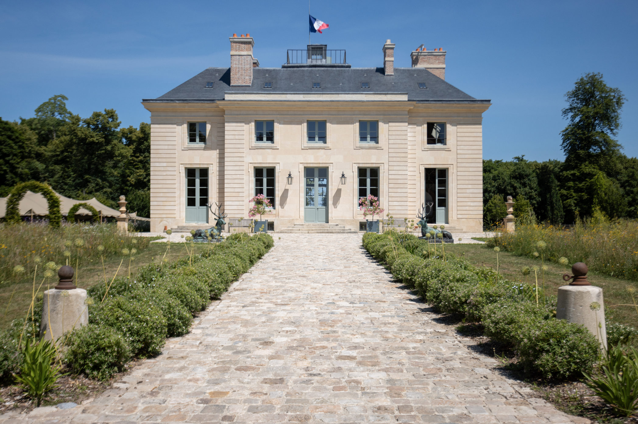 A large, elegant mansion with a symmetrical facade, stone steps, and a French flag flying on top. The building is surrounded by a well-maintained garden with landscape plants and blooming flowers, and a cobblestone pathway leading to the entrance.