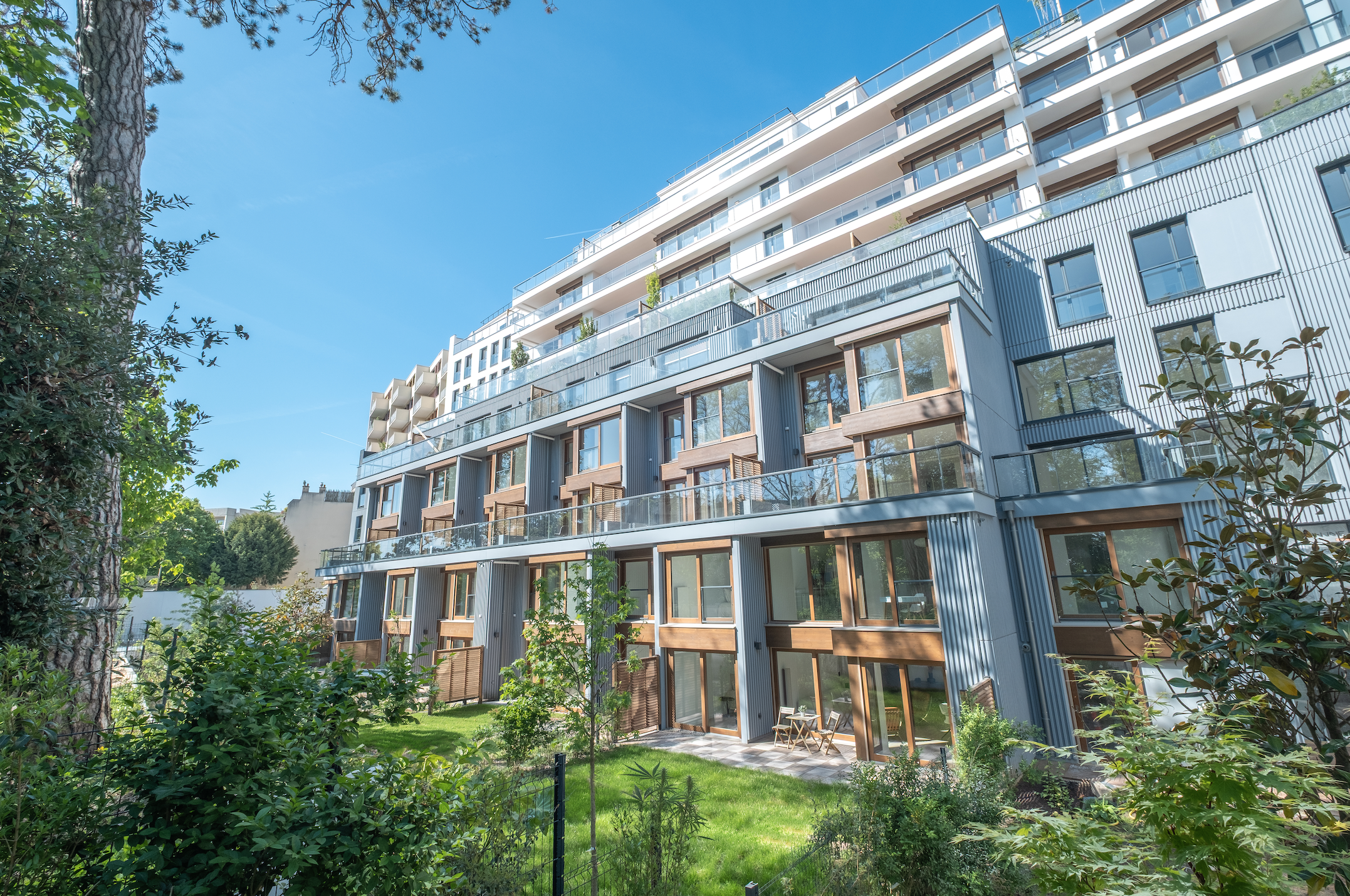 Modern multi-story apartment building with glass balconies, surrounded by greenery, under a clear blue sky.