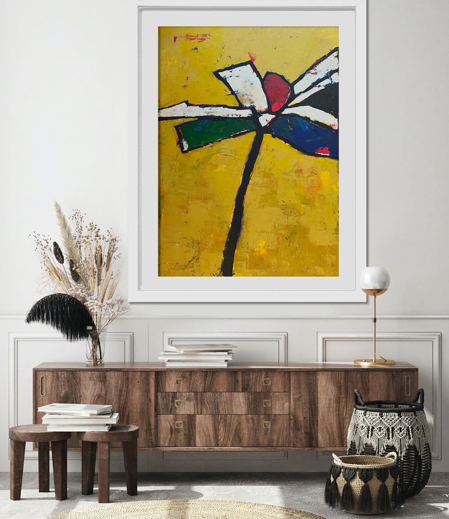 A modern living room with a wooden sideboard, a large abstract painting with bold colors on the wall, decorative dried plants in a vase, a stack of books, woven baskets, and a tripod floor lamp.