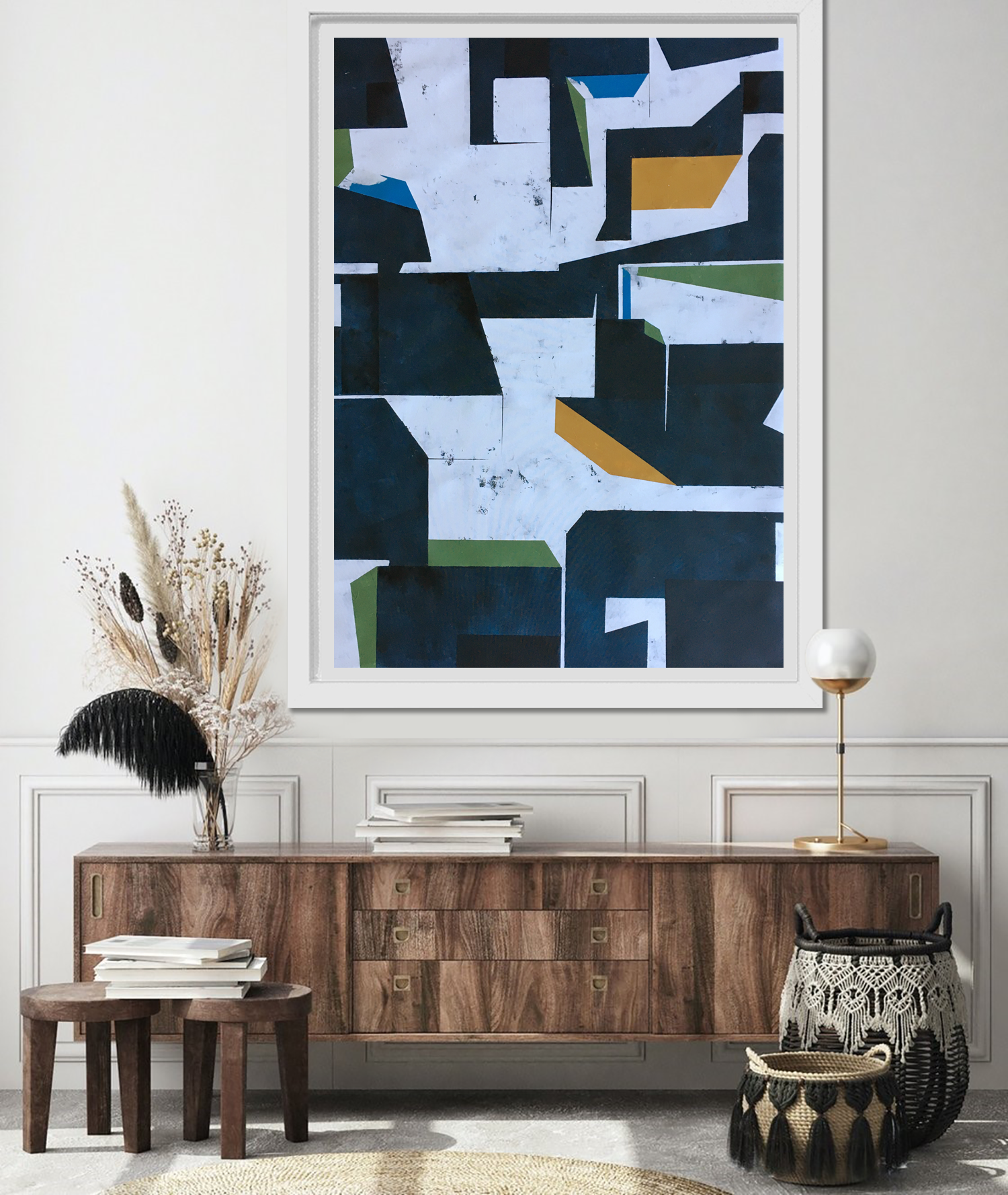 A modern living room decor with a large abstract painting on the wall, a vintage mid-century wooden sideboard, a glass vase with dried plants, stacked books, a black and tan woven basket, and a gold modern table lamp.