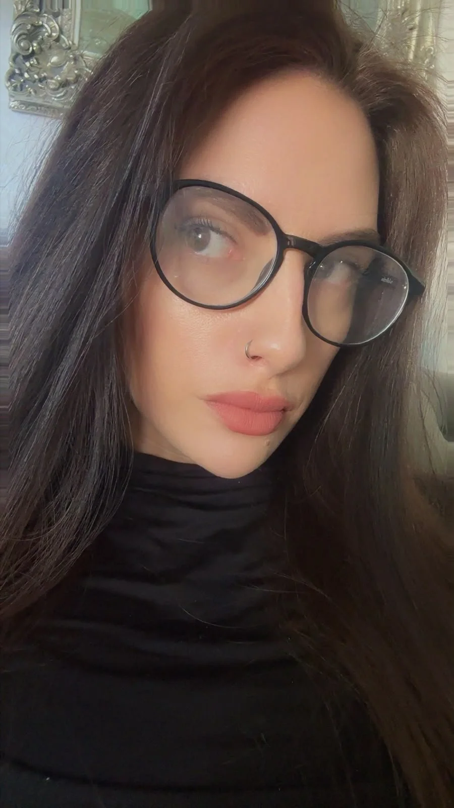 Close-up of a woman with long dark hair, wearing glasses, a nose ring, and a black top.