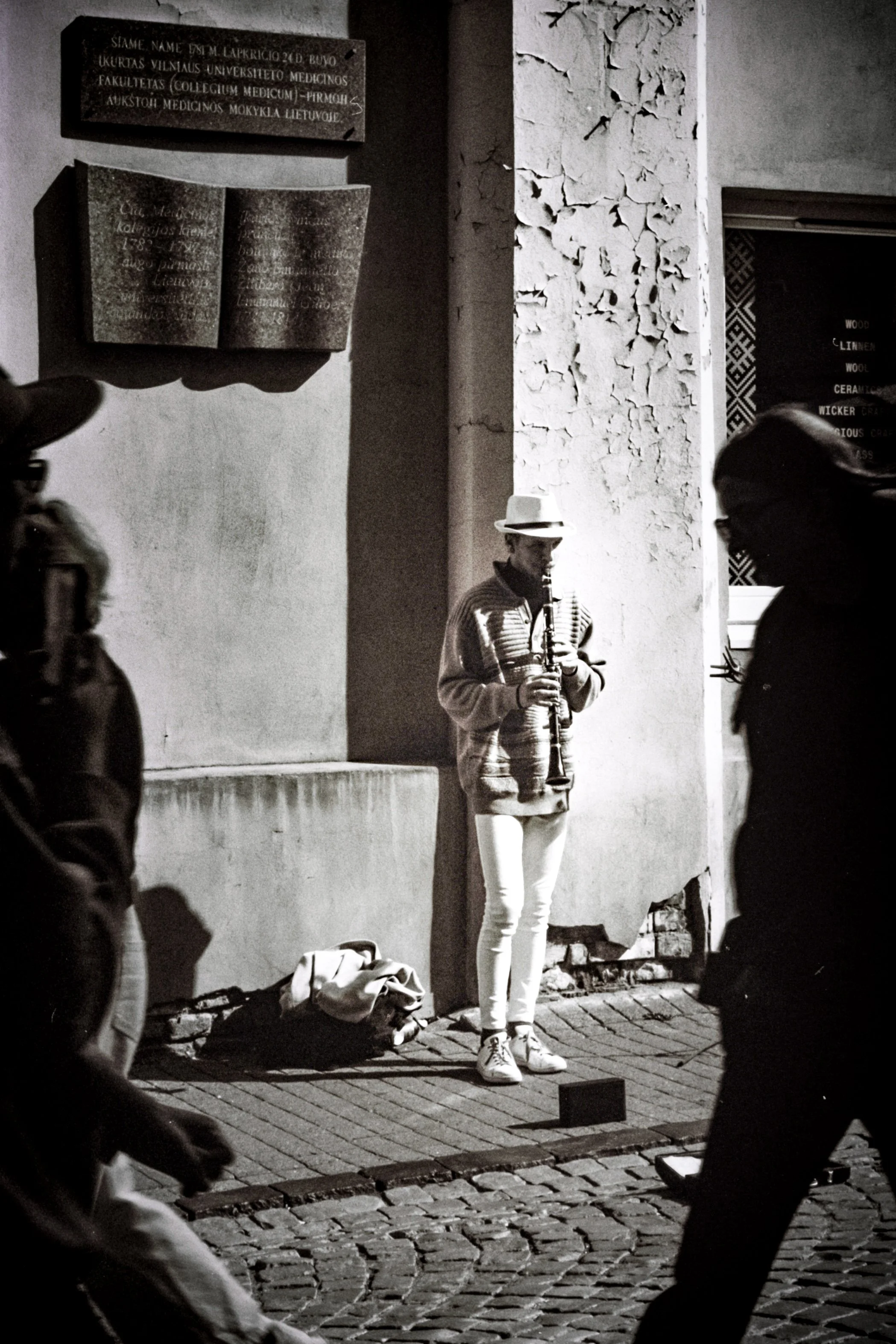 A black and white photo of a street musician playing a clarinet while standing against a textured wall. Several people are walking past, partially visible in the foreground. The musician is wearing a hat, a striped sweater, and white pants.