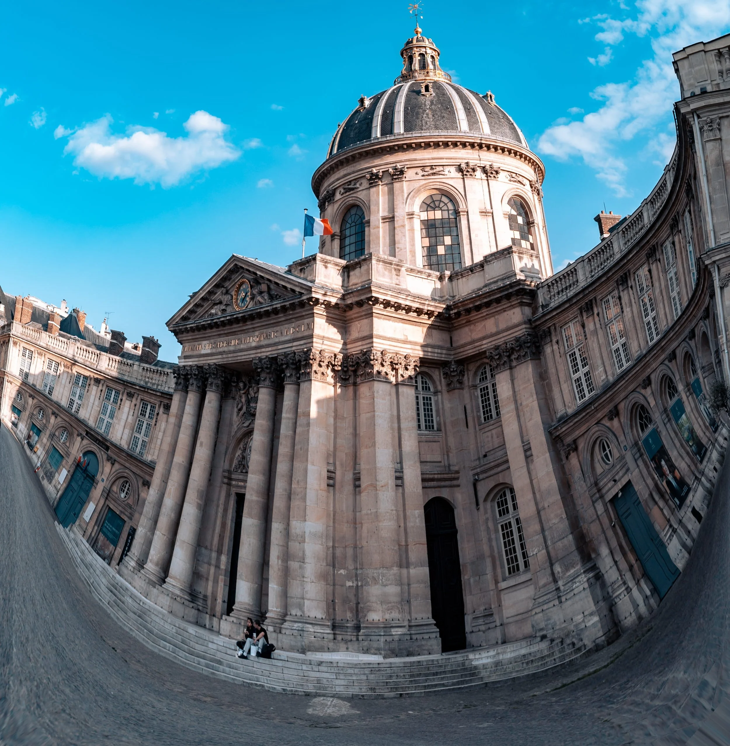 A wide-angle photo of a historic building with a large dome and a French flag, taken from an unusual perspective that distorts the architecture towards the bottom of the image. The building features stone columns and ornate windows, with a partly clo
