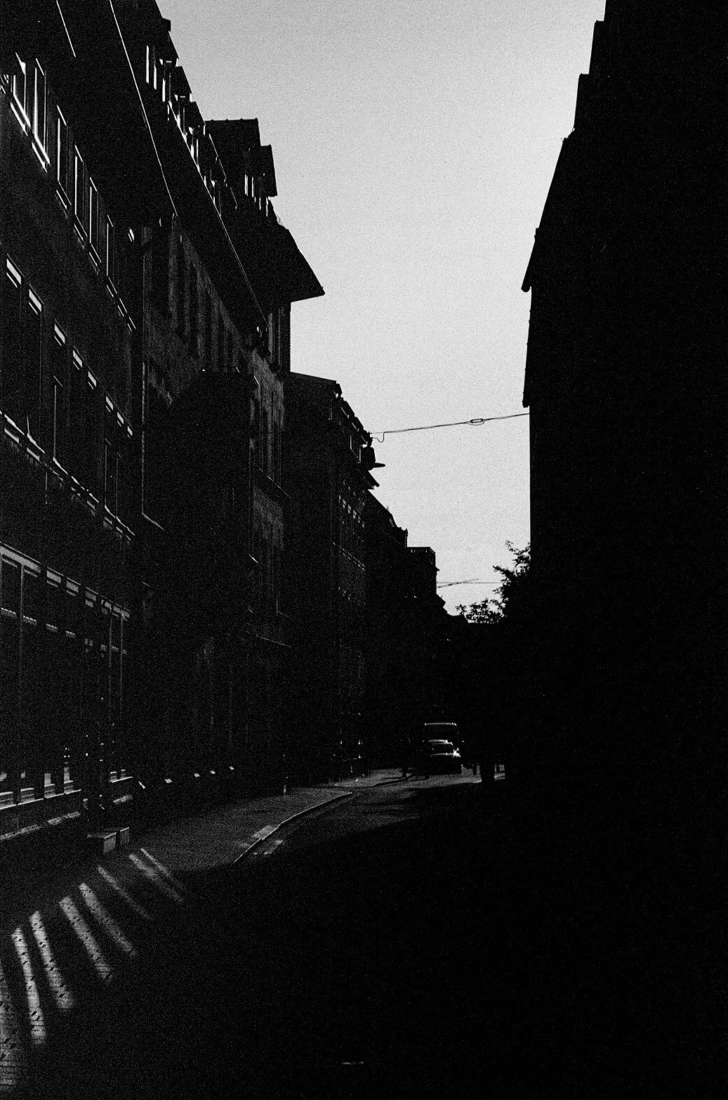 A black-and-white photo of a narrow city street between tall apartment buildings with balconies. The street is mostly in shadows, with some light from the setting or rising sun casting long shadows. There are a few cars parked along the sidewalk and 