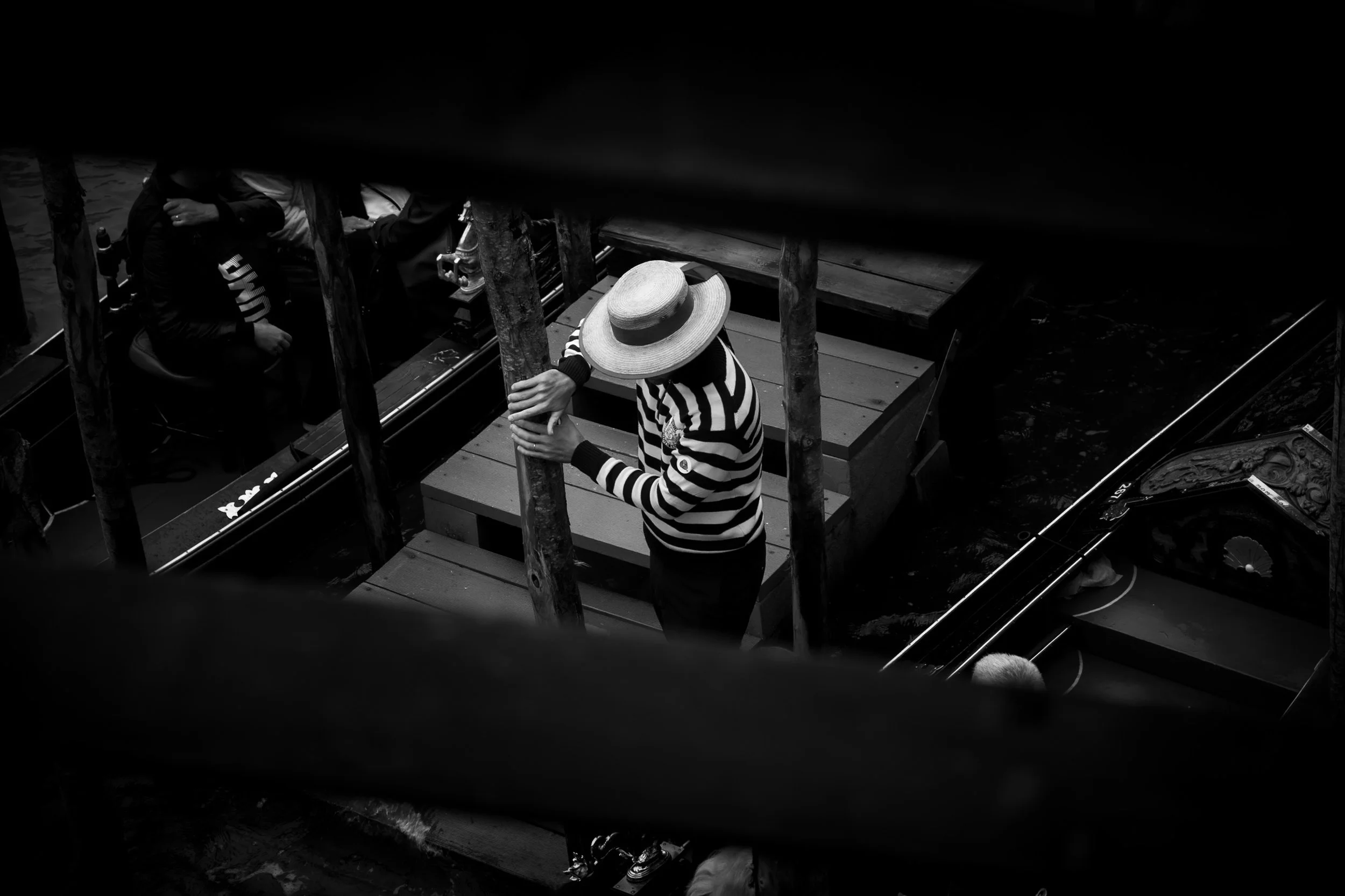A person wearing a striped shirt and wide-brimmed hat leaning on a wooden railing, overlooking water with other people nearby.