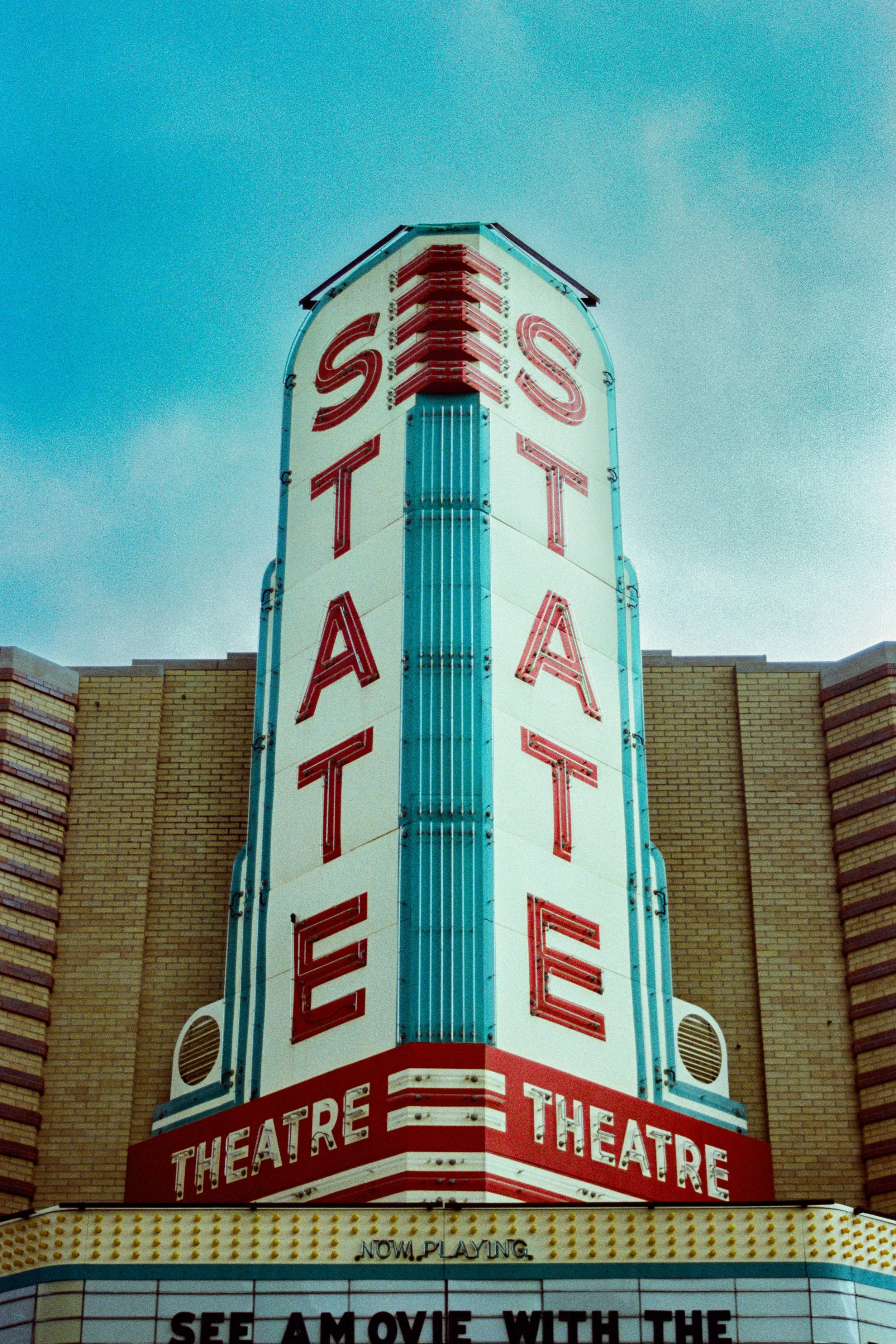The image shows the vertical sign of a movie theater with the word 'THEATRE' in large letters. The sign is retro with illuminated bulbs and red and white colors, set against a brick building with a cloudy sky in the background.