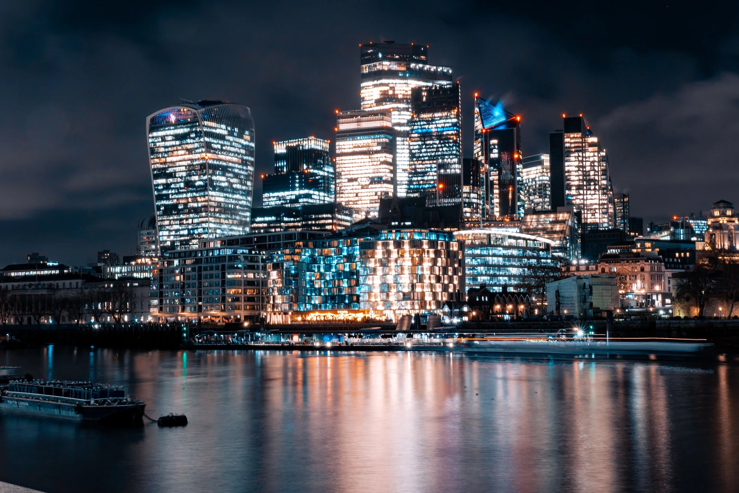 Nighttime city skyline featuring tall illuminated skyscrapers with reflective glass windows, a calm river in the foreground, and dark clouds overhead.