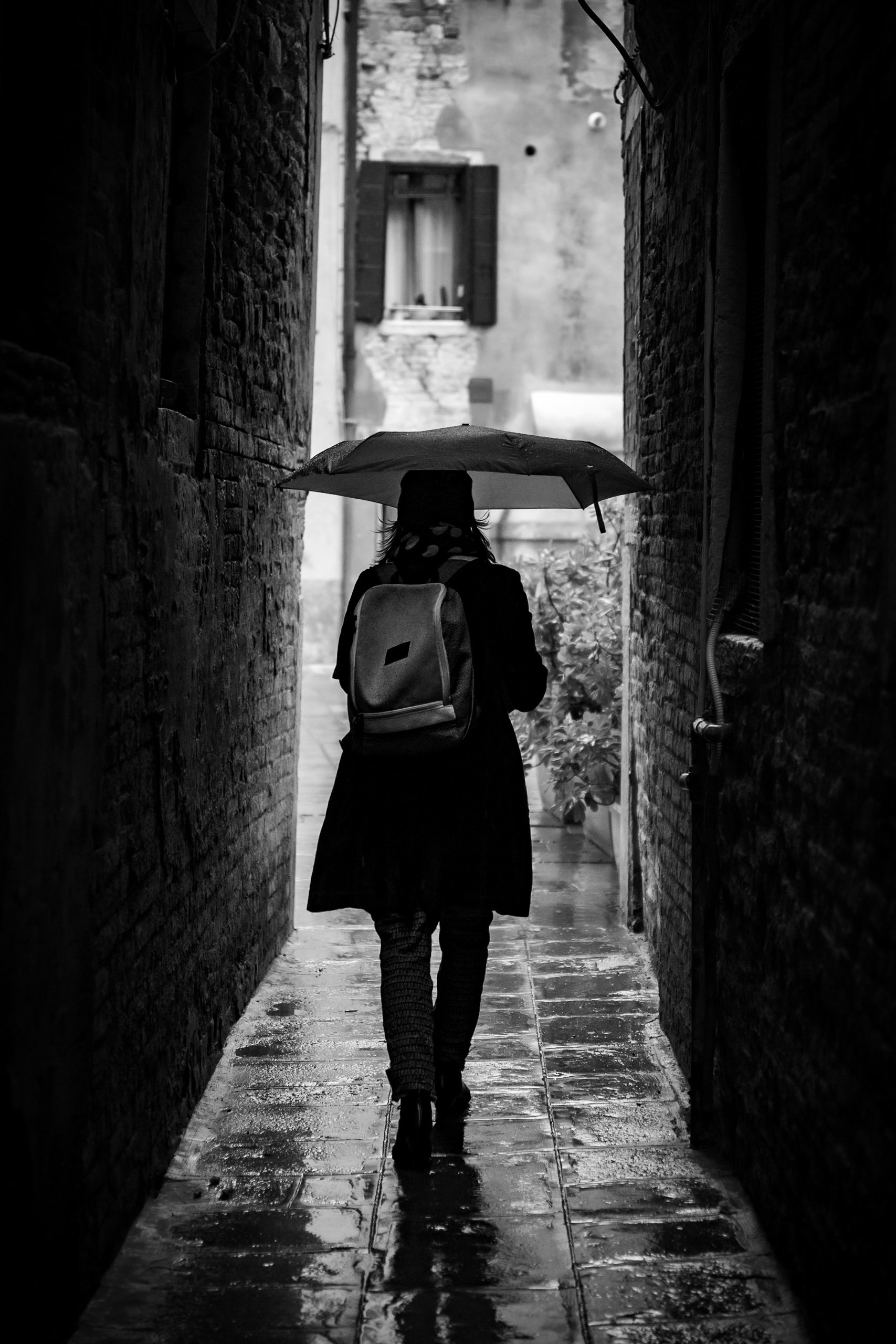 A person walking down a narrow brick alleyway holding an umbrella in the rain, with wet pavement reflecting light.