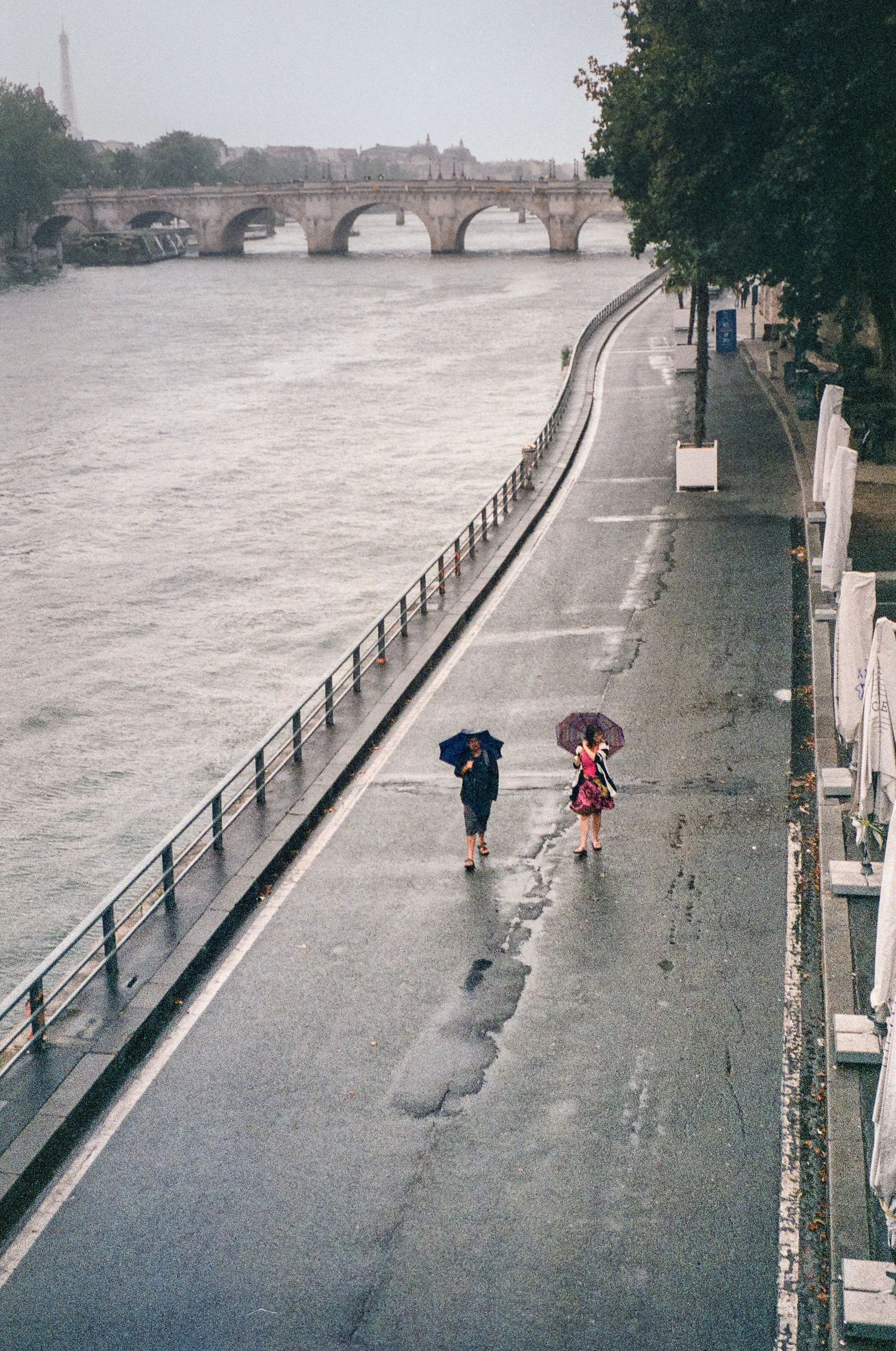 A woman and a girl walking on a wet sidewalk under umbrellas near a river, with a stone bridge in the background on a cloudy day.