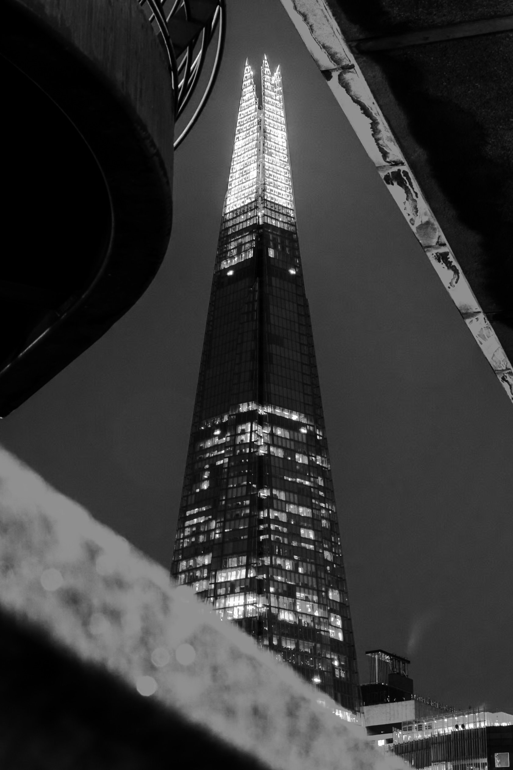 A tall, illuminated modern skyscraper viewed from below at night, with clouds and part of a building visible in the foreground.