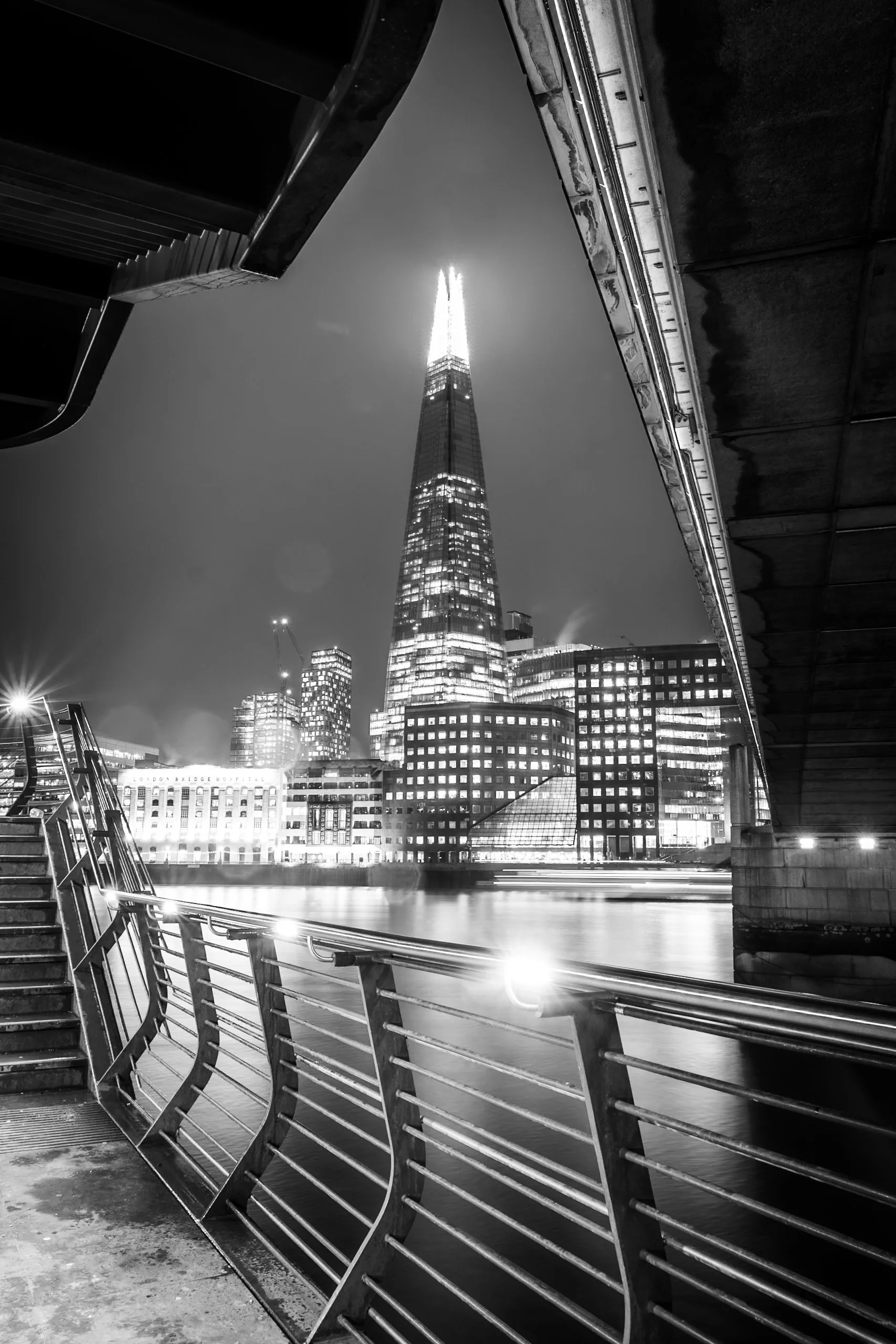 Nighttime cityscape with the illuminated Shard skyscraper and modern buildings behind a metal railing and stairs on a bridge.