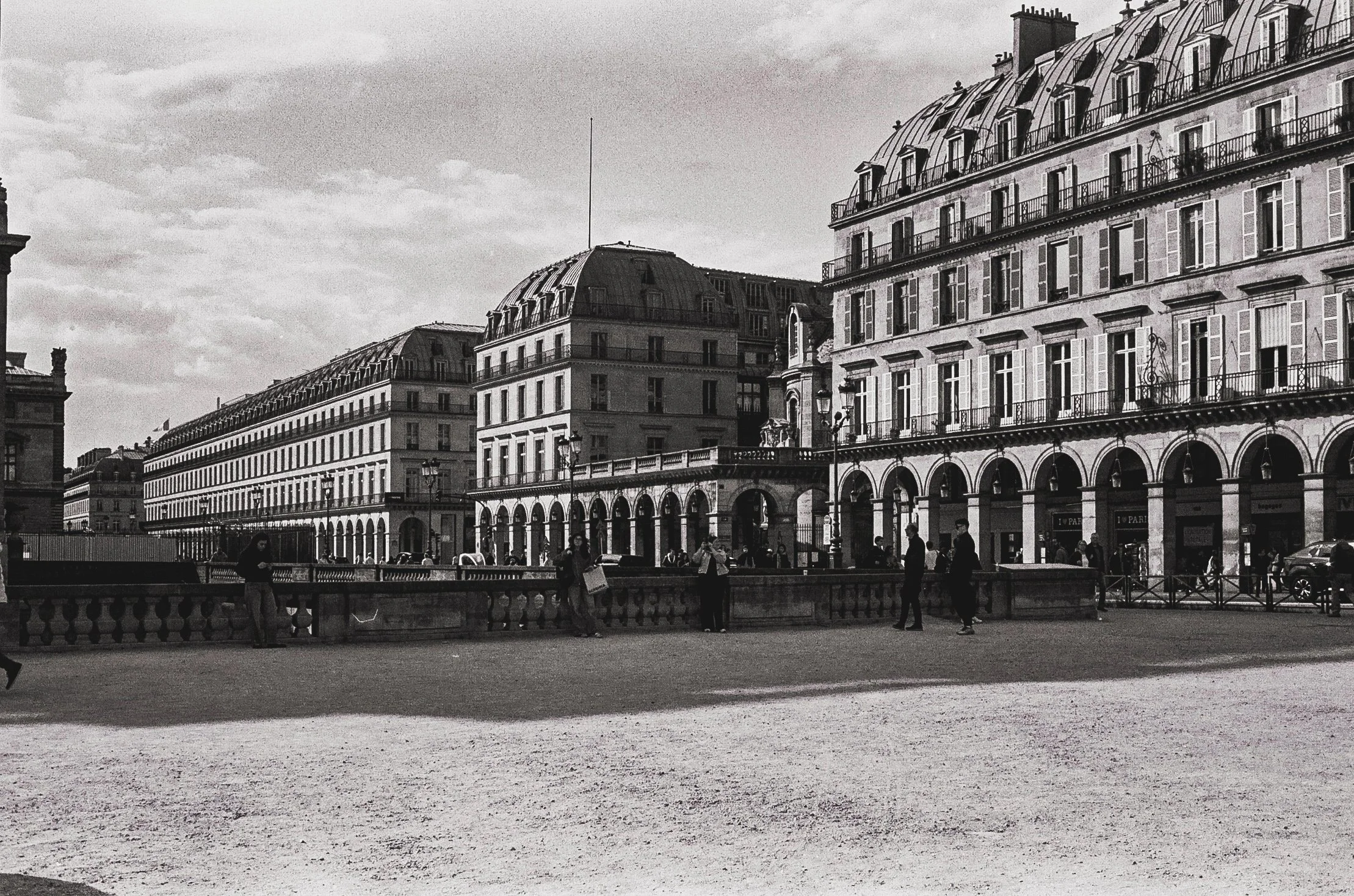 Historic Parisian buildings with arched windows and balconies, seen from a distance under a cloudy sky, with pedestrians walking along a bridge in the foreground.