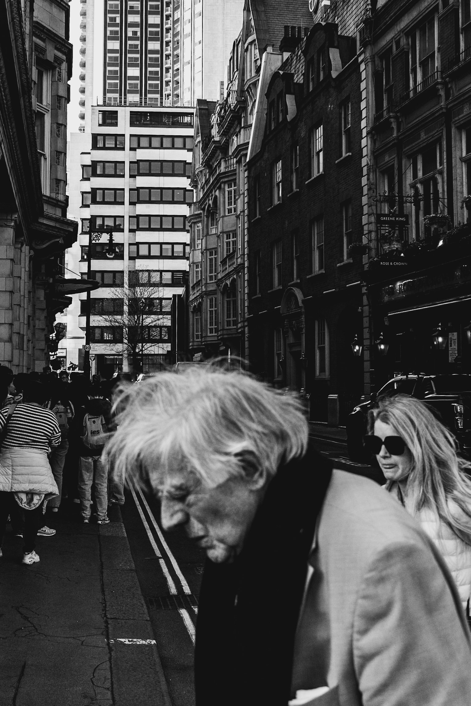 Black and white photo showing a busy city street filled with pedestrians, tall buildings, and storefronts. A woman with sunglasses in the foreground looks to the side.
