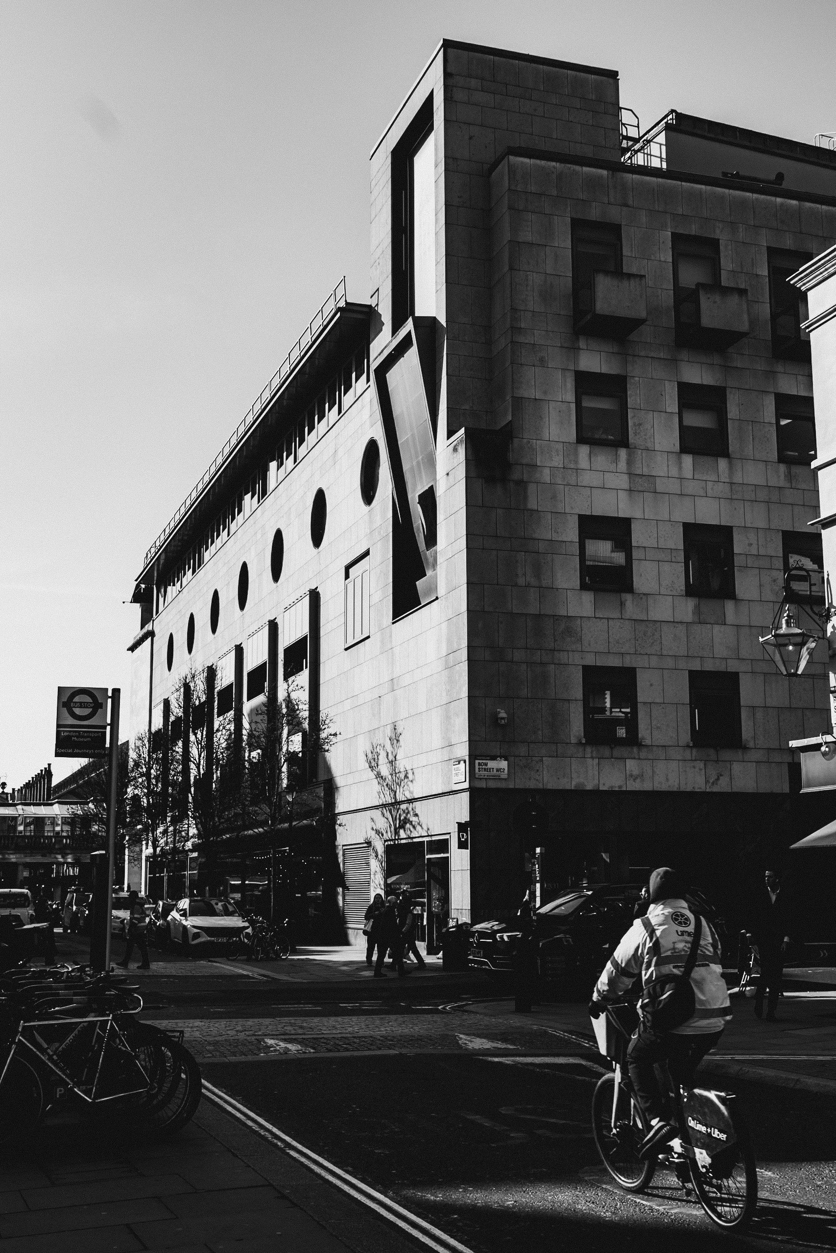 Street scene in an urban area with a modern building, bicycles parked, and a cyclist riding a bike labeled Uber Eats. Pedestrians are visible, and the sky is clear.