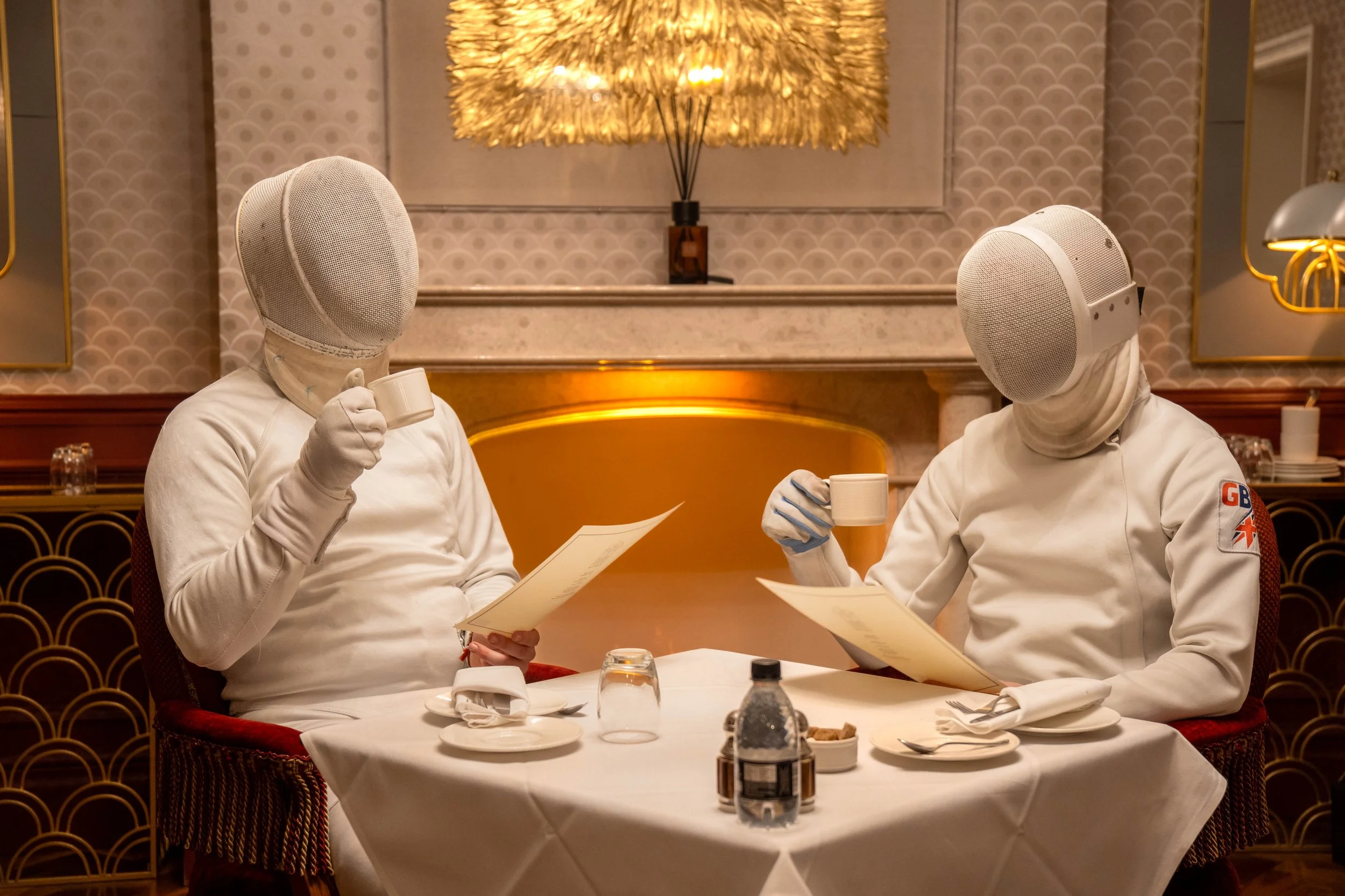 Two fencers in fencing gear sitting at a restaurant table, drinking coffee and holding menus, with a lit decorative lamp in the background.