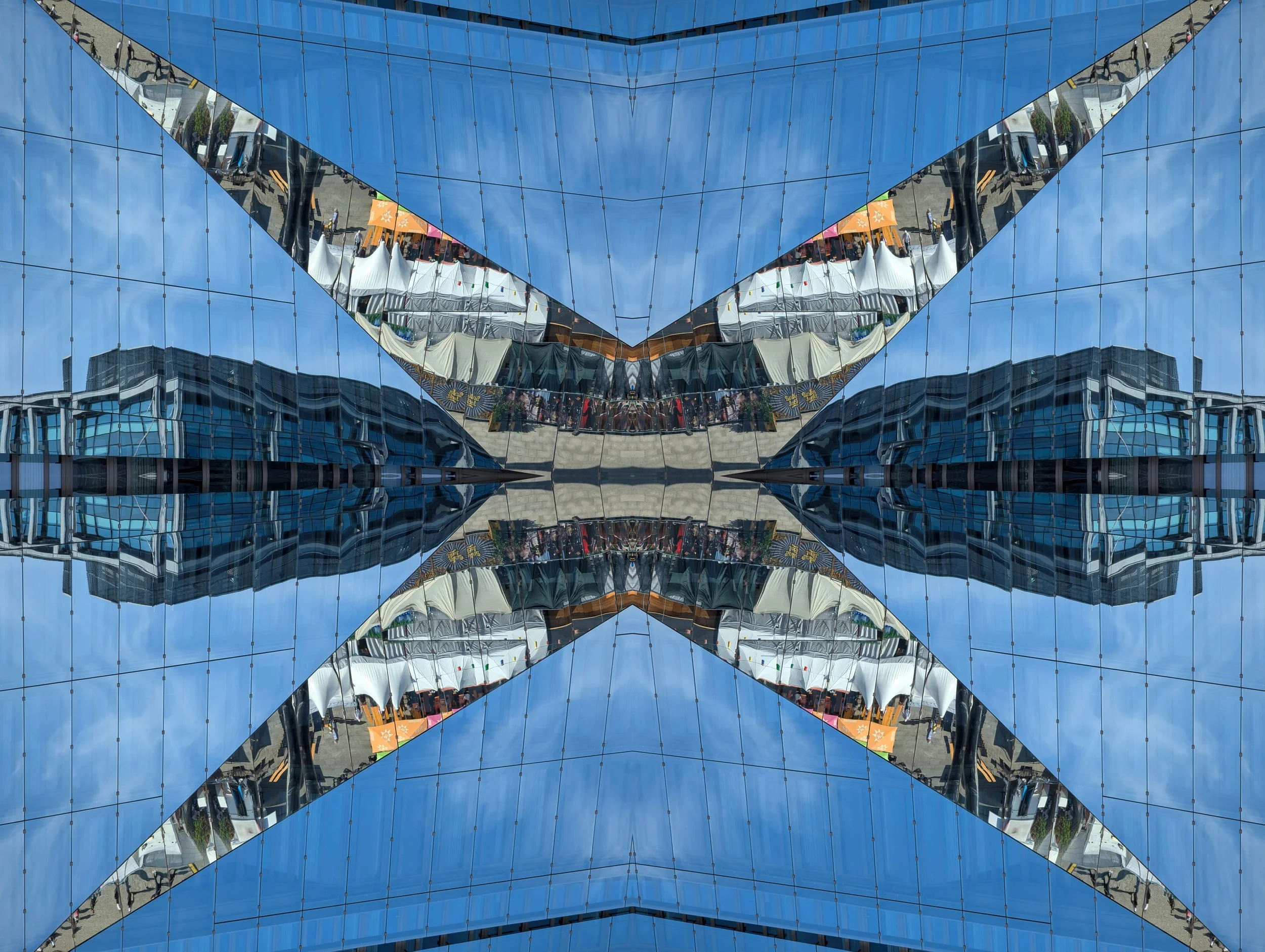 A reflected view of tents and people at an outdoor event, captured in a building's mirrored glass facade that creates an abstract, symmetrical pattern.