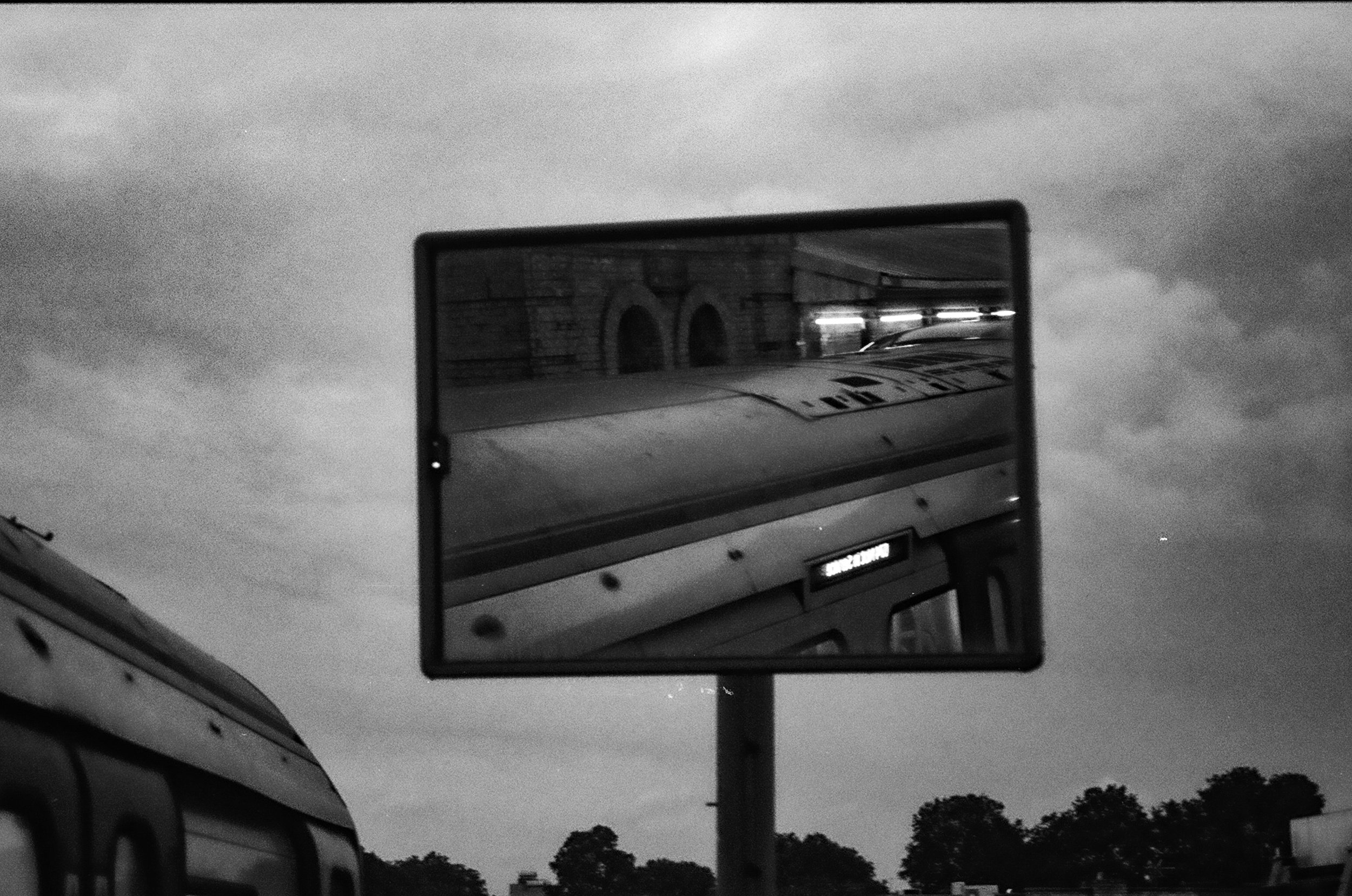 Black and white photo of a road sign and mirror on a vehicle. The road sign is in focus, with trees and cloudy sky in the background.