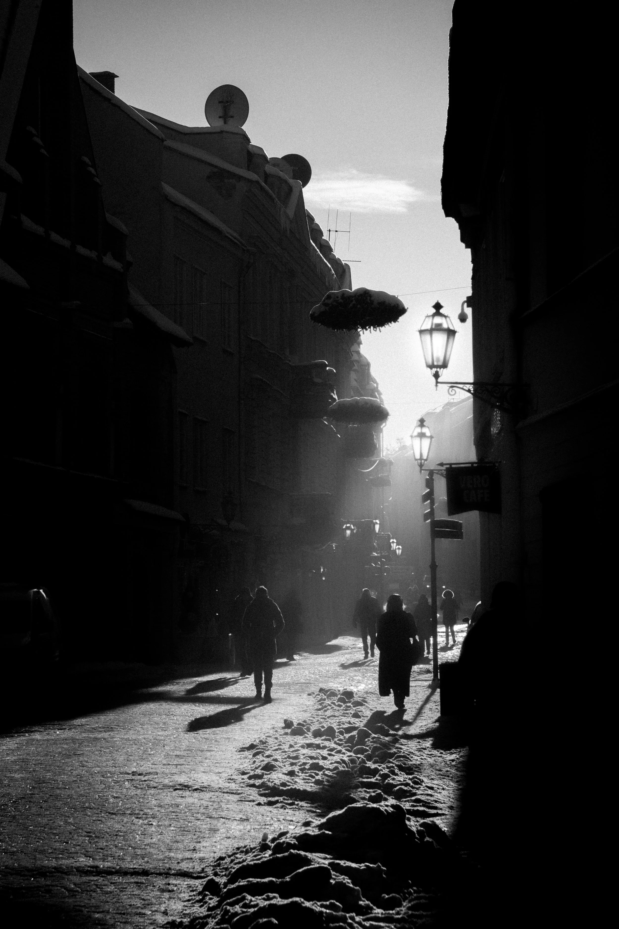 People walking on a snowy street with shadows cast by the setting or rising sun, buildings with satellite dishes and street lamps visible, black and white photo.