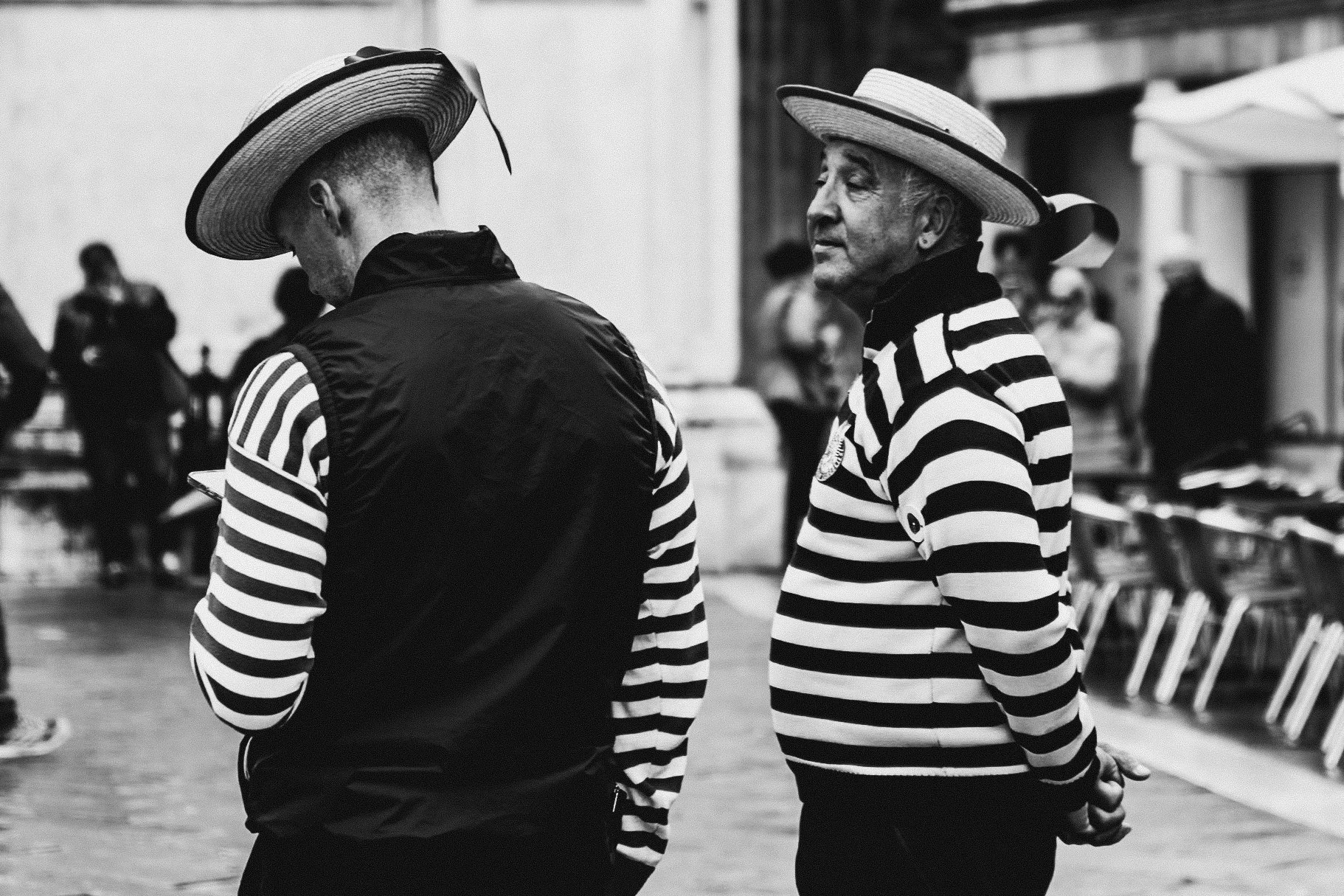 Two women in striped shirts and wide-brimmed hats standing in a public setting with chairs and other people in the background.