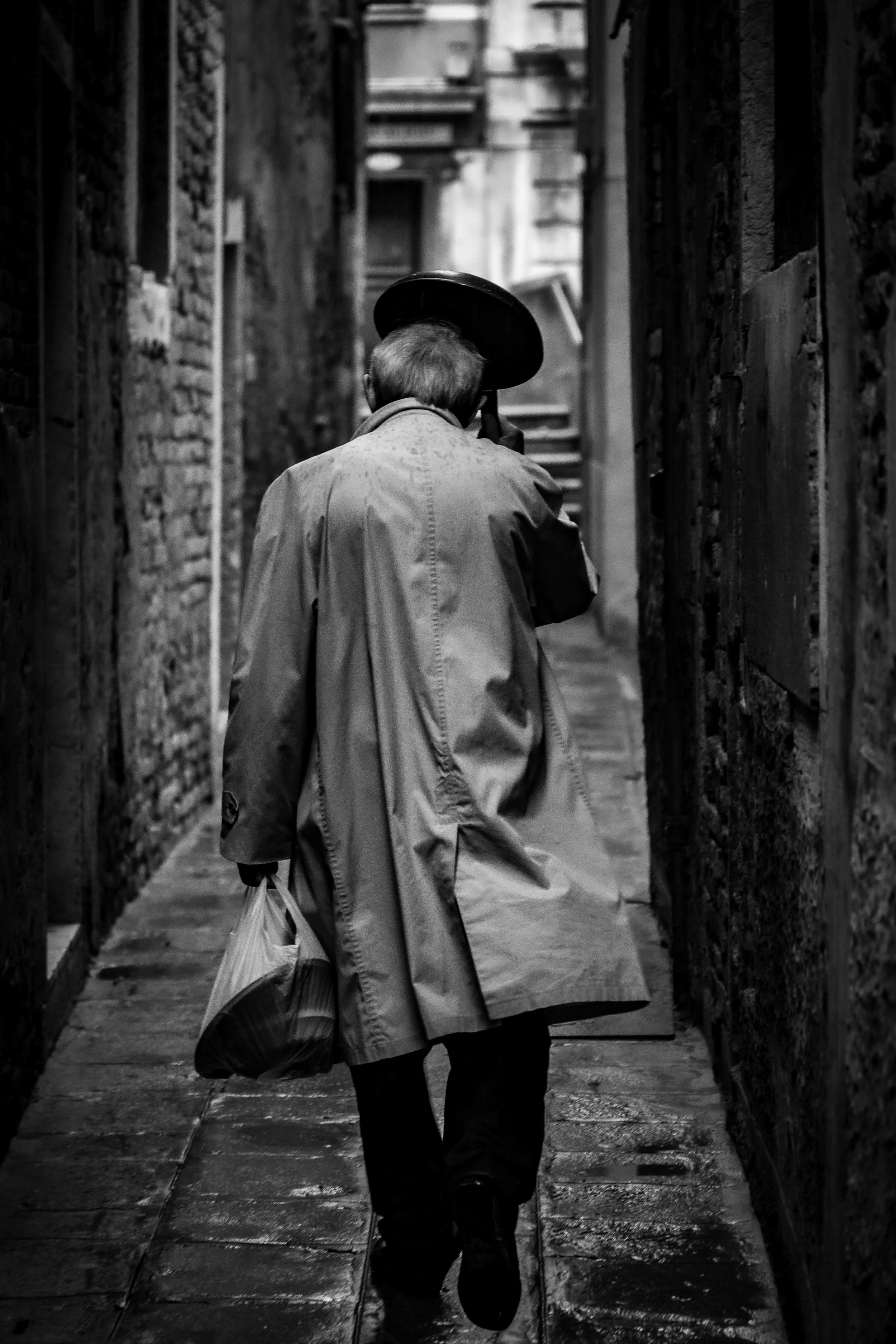 A black-and-white photograph of a person walking down a narrow alleyway with brick walls on either side, holding a hat in their right hand and a bag in their left, with a staircase in the background.
