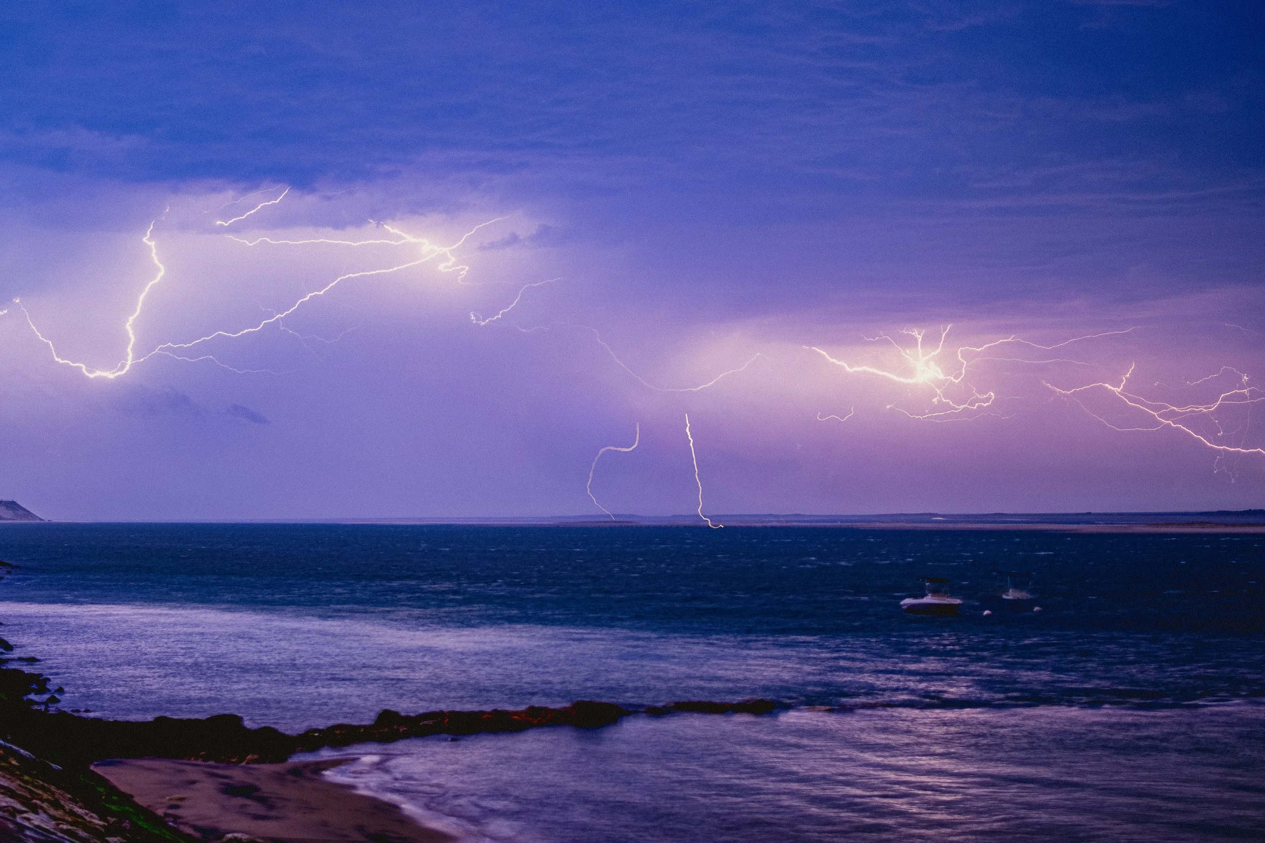 Lightning strikes over a body of water during a thunderstorm at night, with a cloudy sky and a distant shoreline.