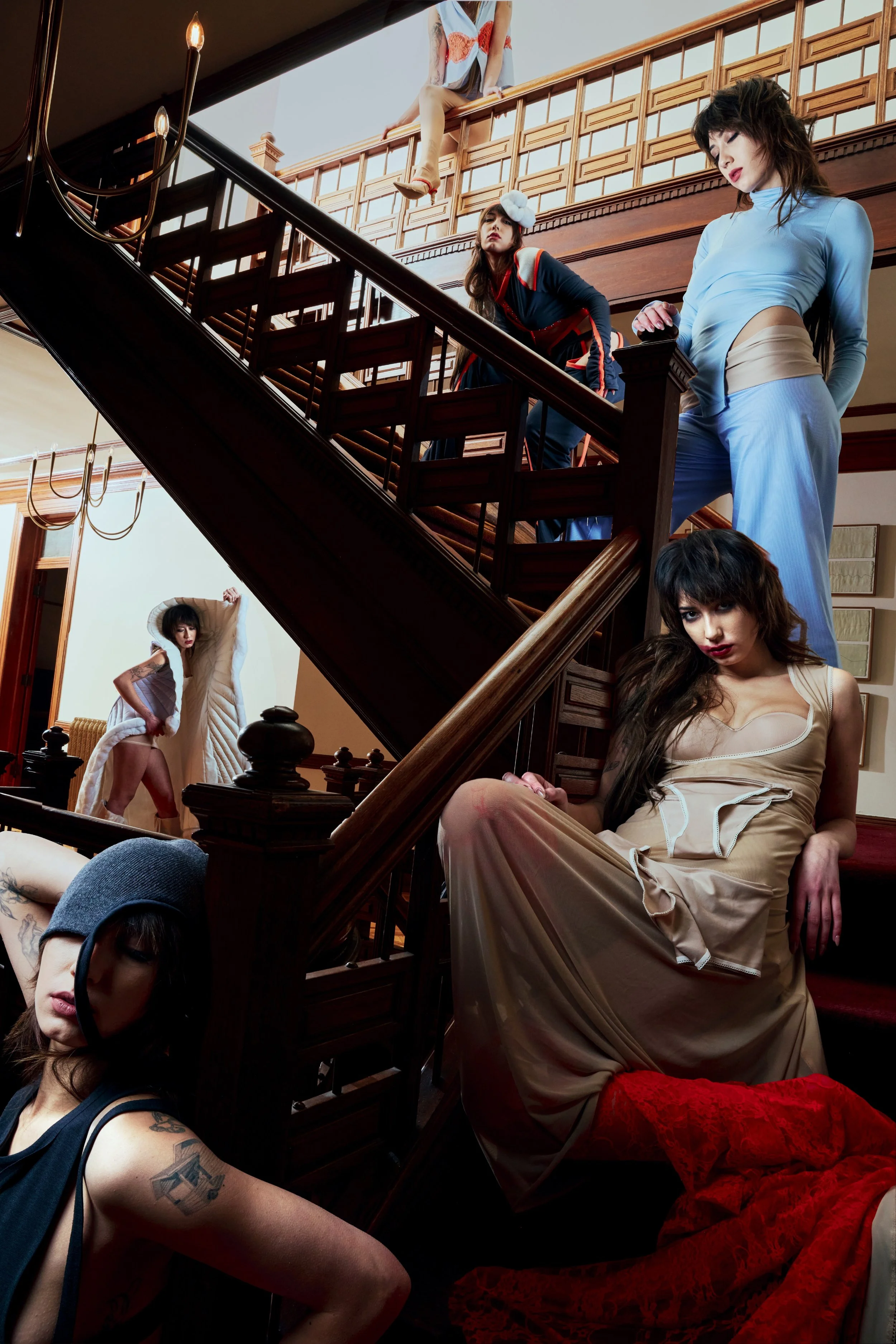 Multiple women with tattoos and vintage clothing sit and pose on a grand wooden staircase inside a well-lit room with large windows and chandeliers, creating an artistic and edgy scene.