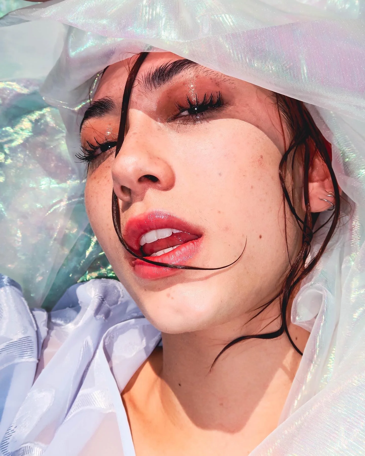 A close-up of a woman's face with makeup, wet hair, and iridescent fabric around her head, lying on a holographic surface.