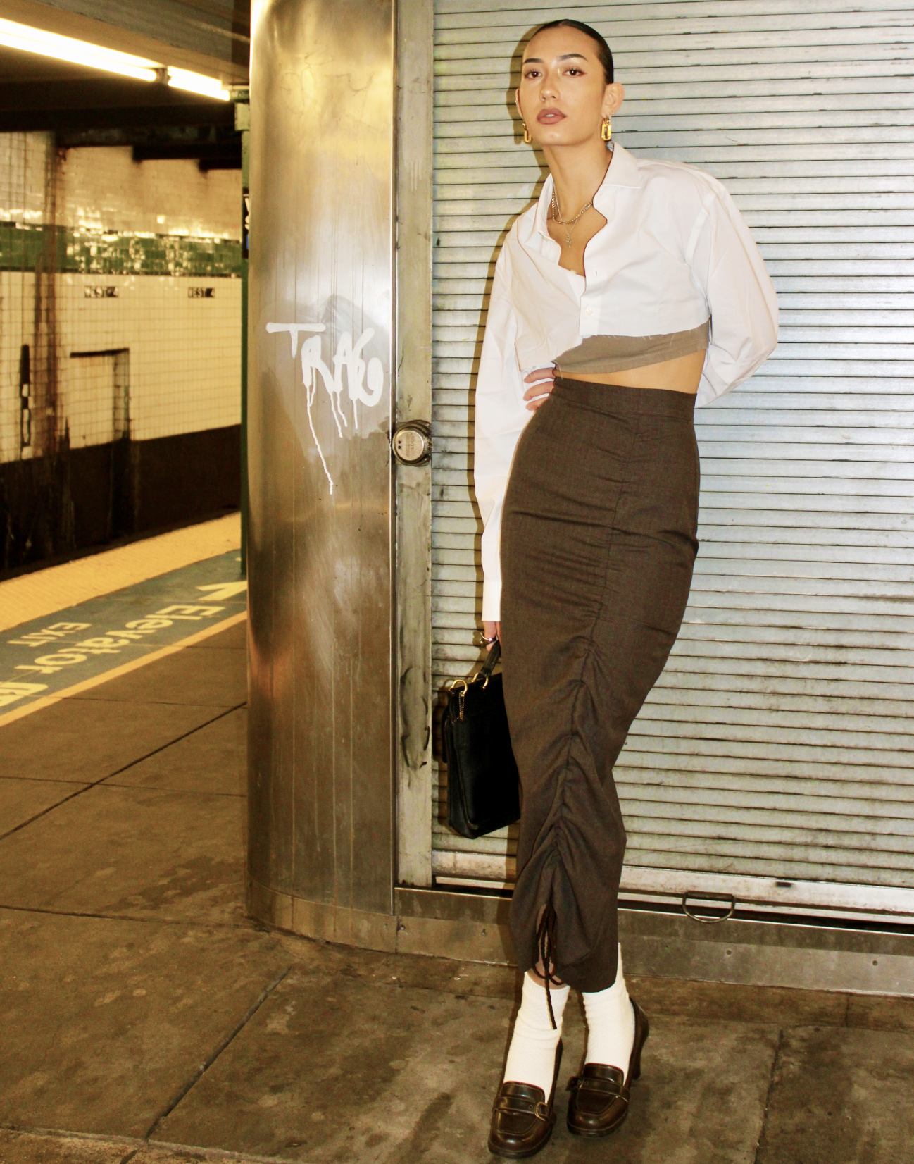 A fashionable young woman standing in front of a metal shop shutter, wearing a white shirt with part of it pulled up to reveal a beige underlayer, a high-waisted dark skirt, white socks, and black loafers. She has gold jewelry and is holding a black handbag.