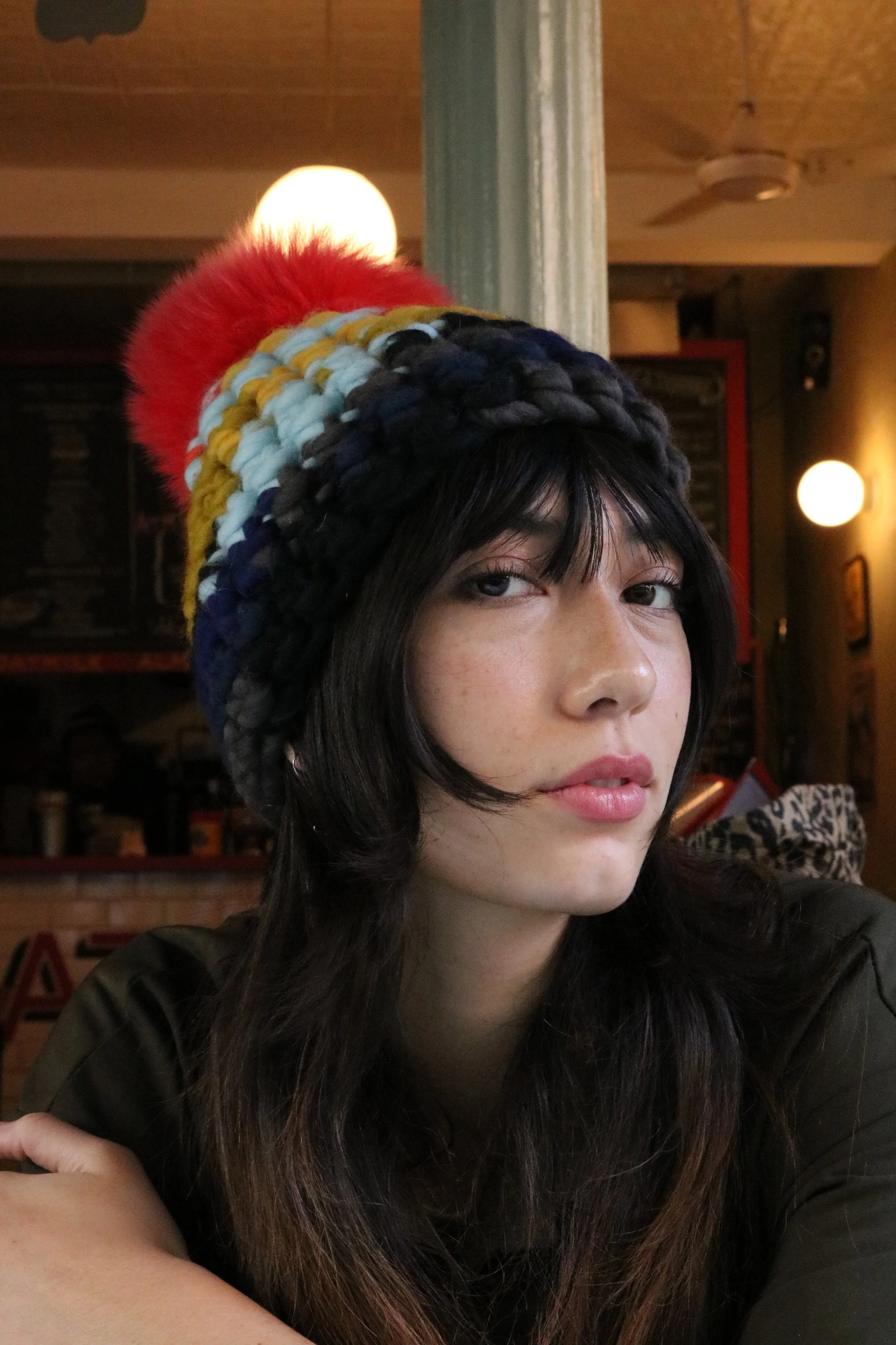 A woman with dark hair wearing a multicolored knitted hat with red, yellow, light blue, and black sections, in a dimly lit room with warm lighting and ceiling fans.