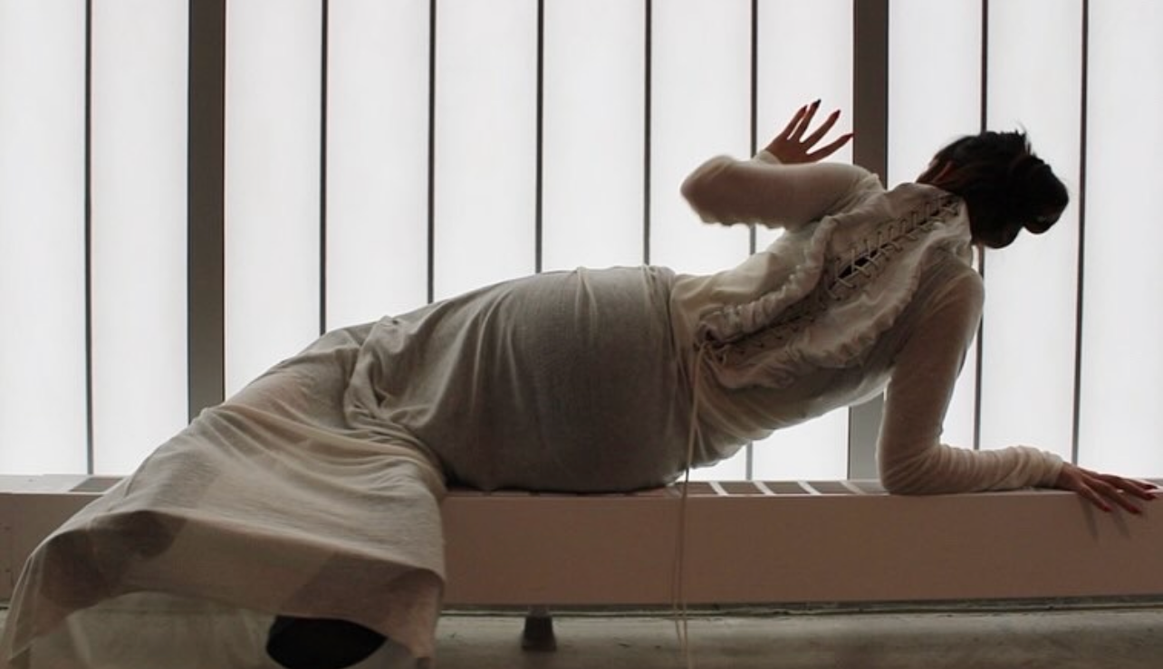 A woman is lying on her side on a low surface, wearing a long beige dress and a light-colored shawl, with her hand touching a vertical window with bars in the background.