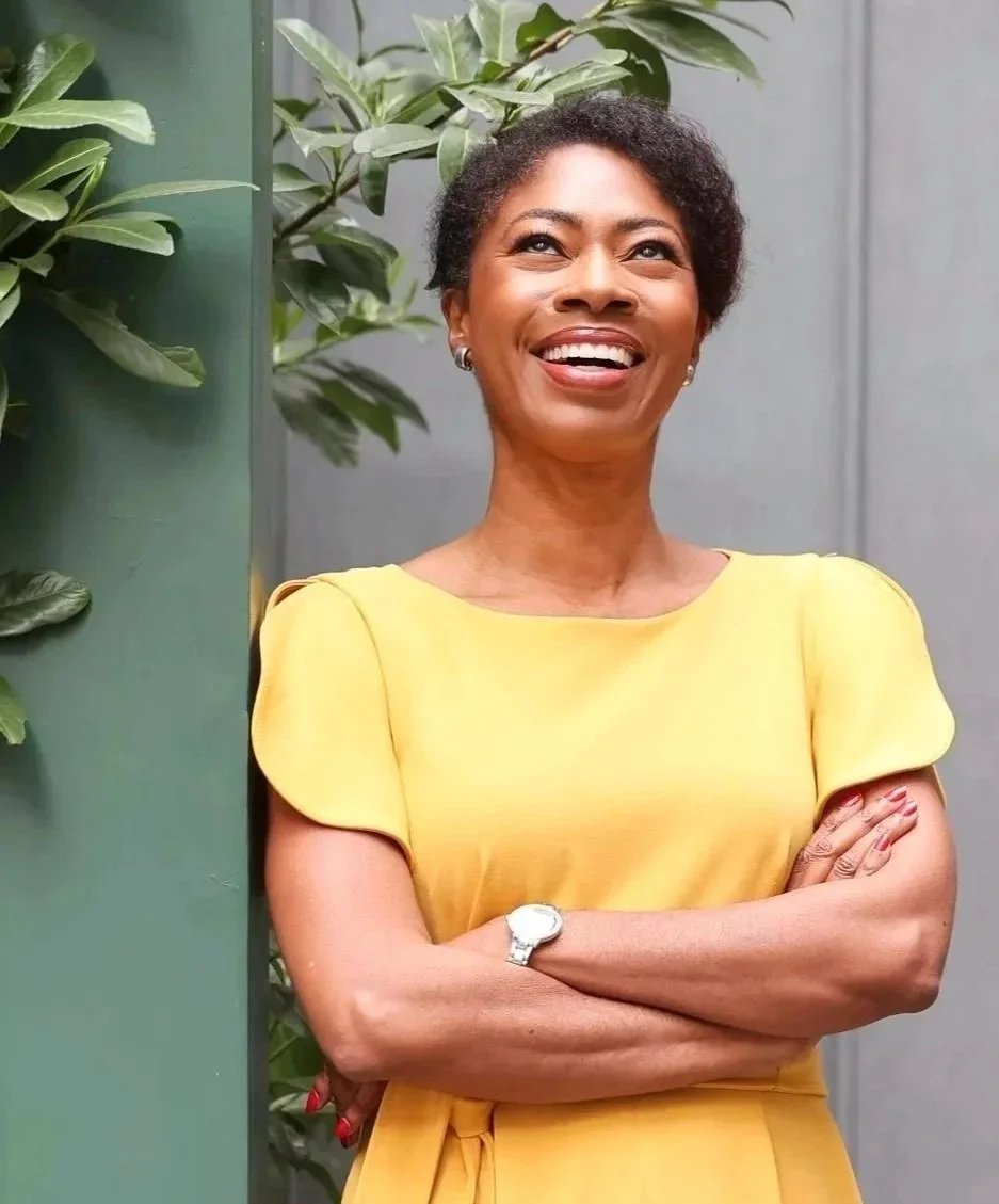 A woman with short curly hair, wearing a yellow dress, smiling, with arms crossed, standing outdoors near green plants and a gray wall.