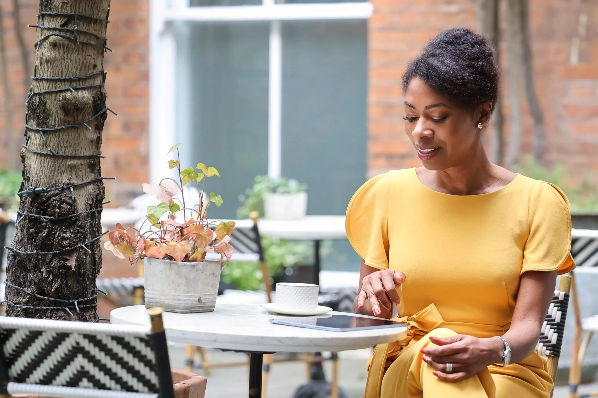 A woman in a yellow dress sitting at an outdoor table, looking down with a slight smile. There are plants and outdoor furniture around her, and a tree trunk wrapped with string lights on the left side of the image.
