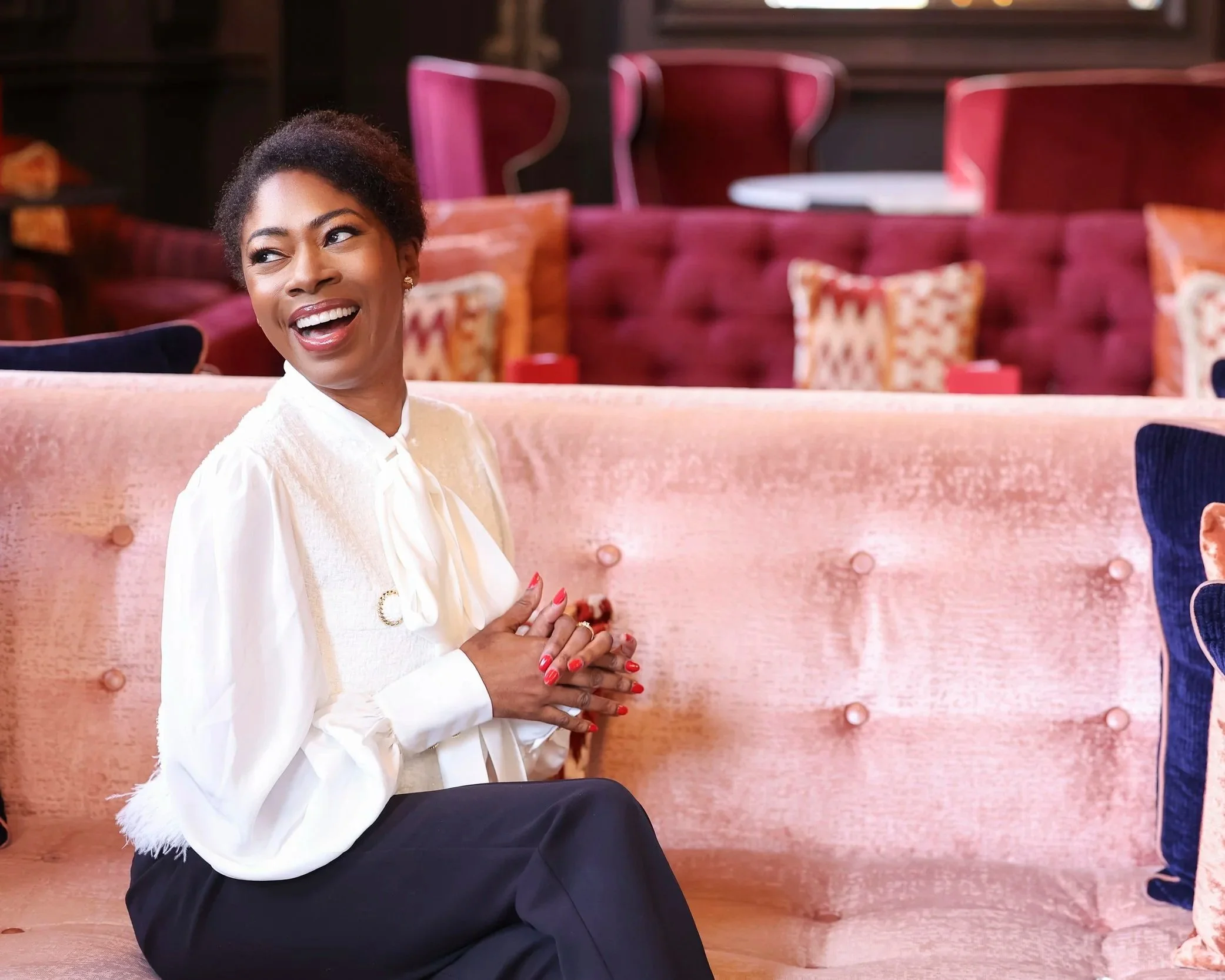 A woman with short dark hair, smiling with her hands clasped together, sitting on a pink upholstered bench in a room with pink and purple chairs and pillows in the background.