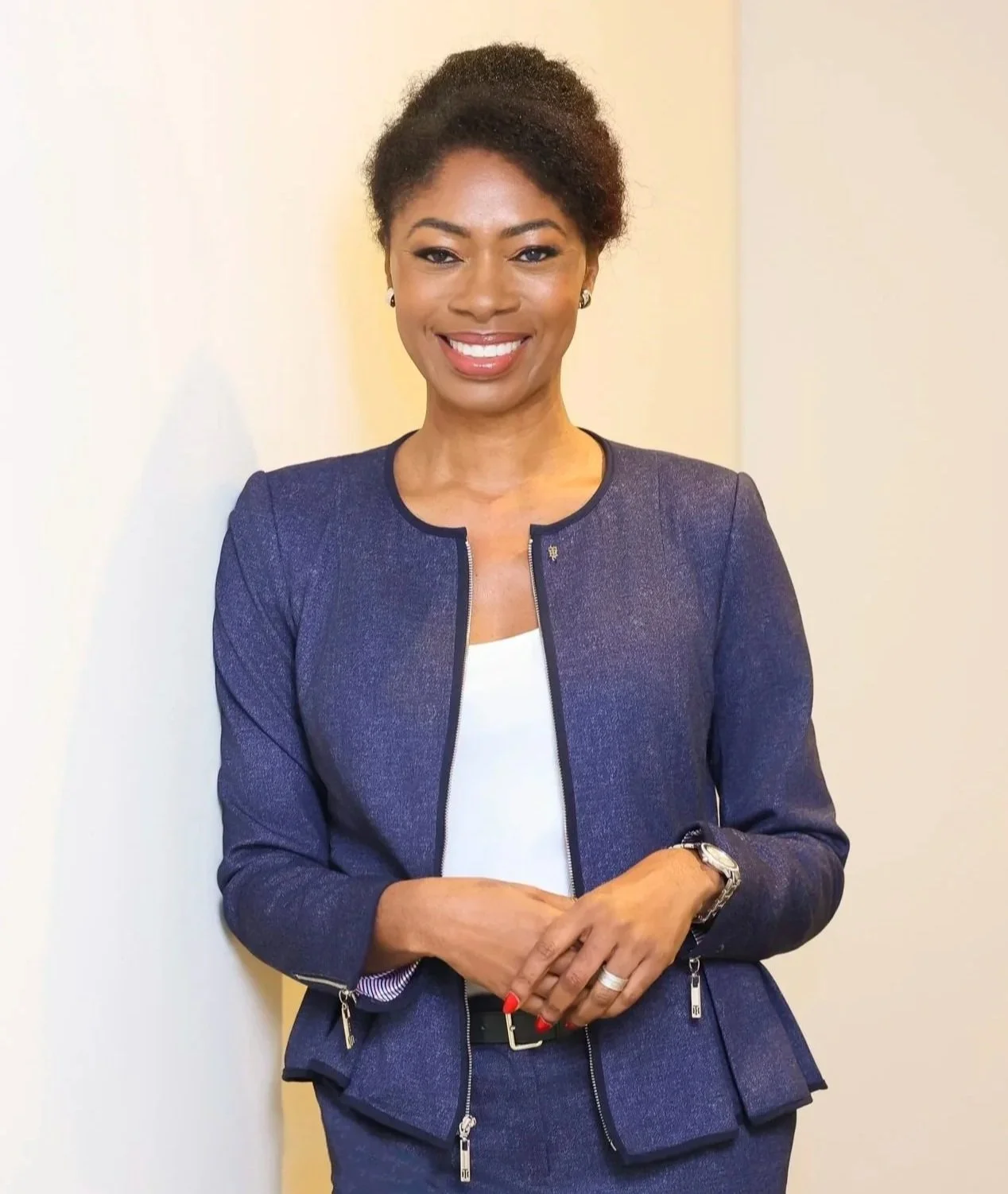 A woman in a dark blue blazer and skirt, smiling and posing against a plain beige wall.