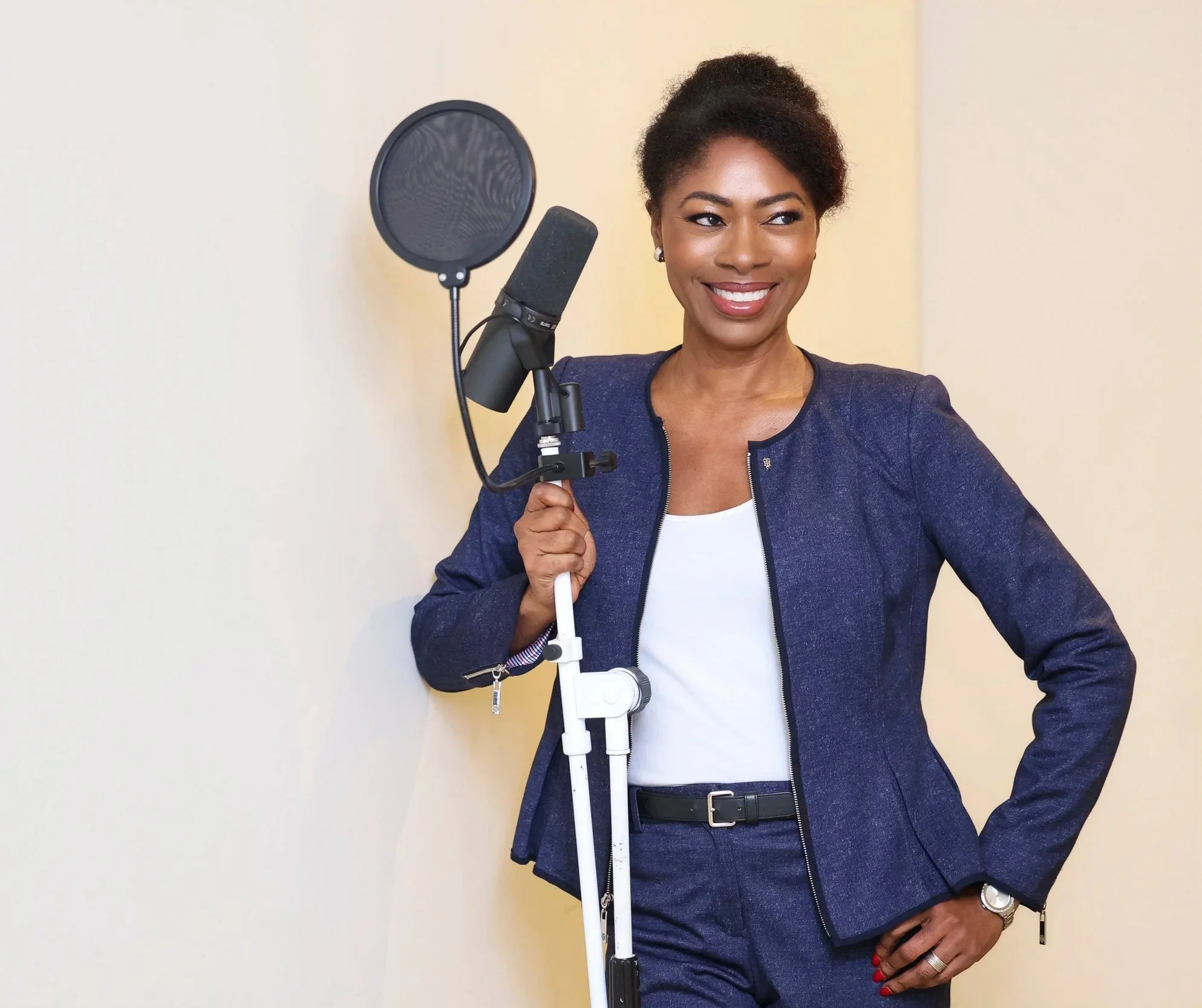 A woman with dark skin smiling in front of a plain wall, holding a microphone stand with a microphone attached.