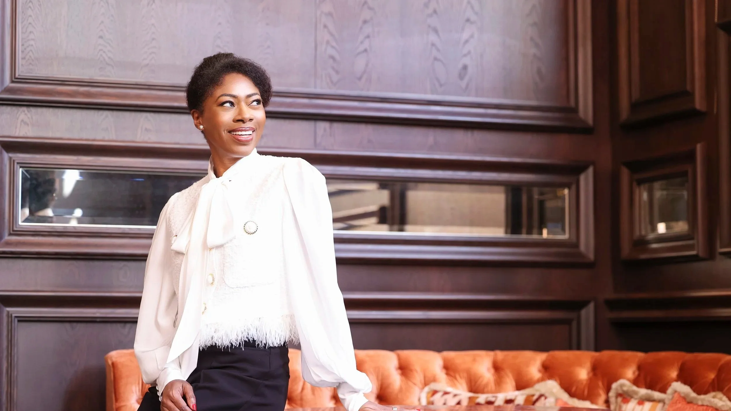 A woman with short curly hair and a white blazer smiling while standing in a room with dark wooden paneling and an orange tufted sofa.