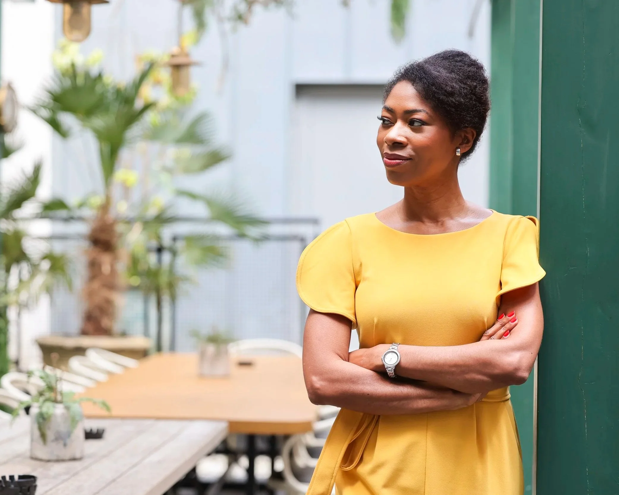 A woman standing outdoors with her arms crossed, wearing a yellow dress. She has short, curly black hair and is wearing a silver watch and earrings. The background includes green plants and a modern building.