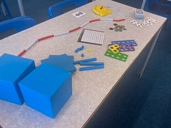 Educational materials on a table including large blue blocks, colored pattern blocks, plastic connecting rods, a yellow calculator, a coin pile, dice, a sheet of paper, and a white device with a screen.