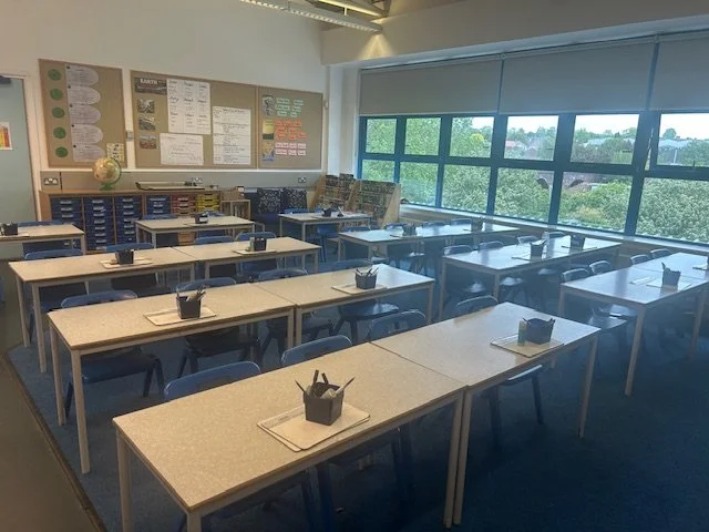 Empty classroom with organized desks, chairs, educational materials, and large window with a view of trees.