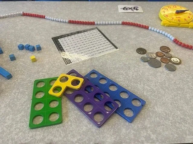 Educational materials including colored counting blocks, a math grid, a bead string, a measuring tape, and coins on a light gray surface.