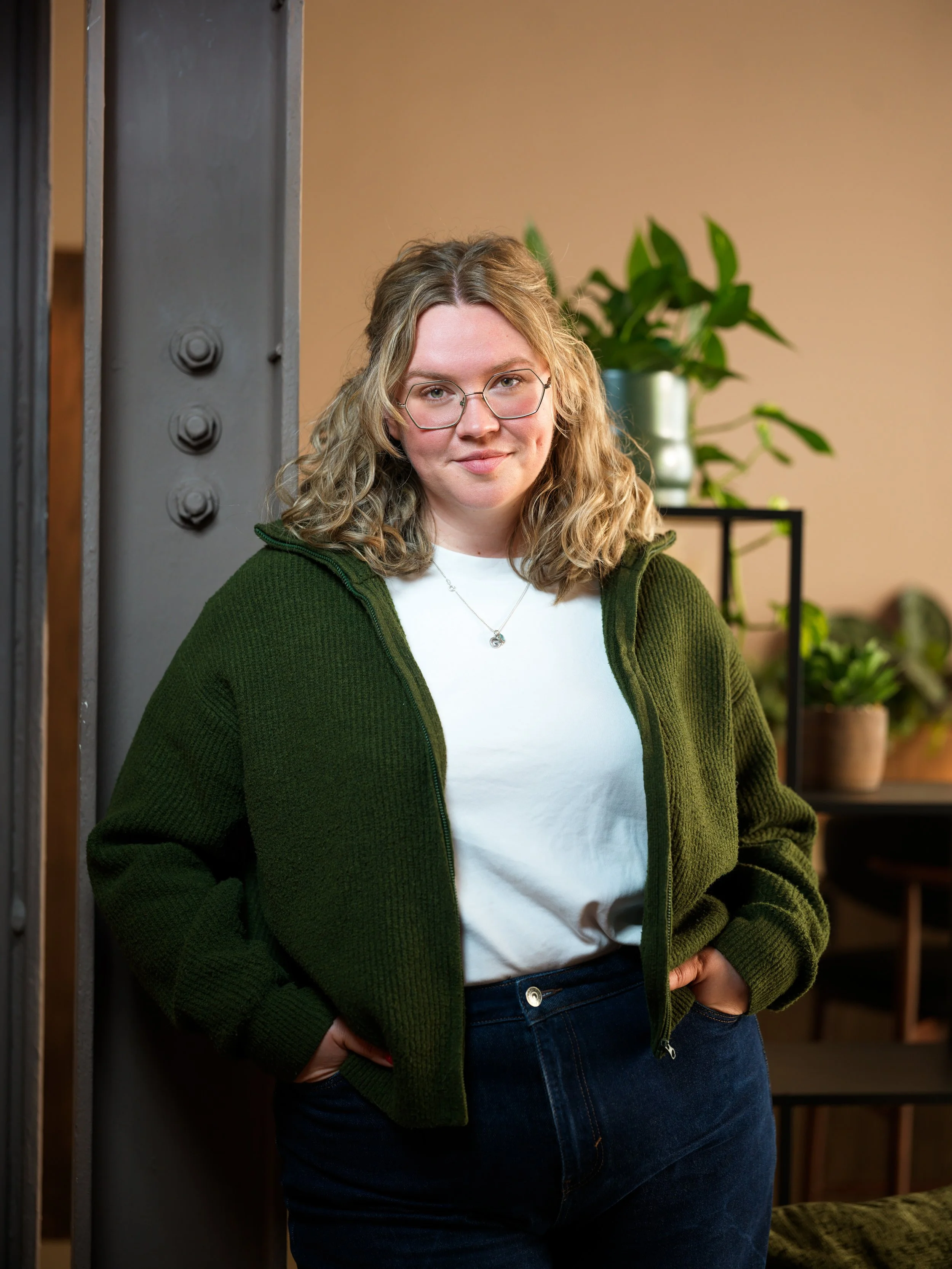 A woman with blonde, curly hair wearing glasses, a white shirt, green zip-up jacket, and blue jeans posing indoors with a background of houseplants and shelving.